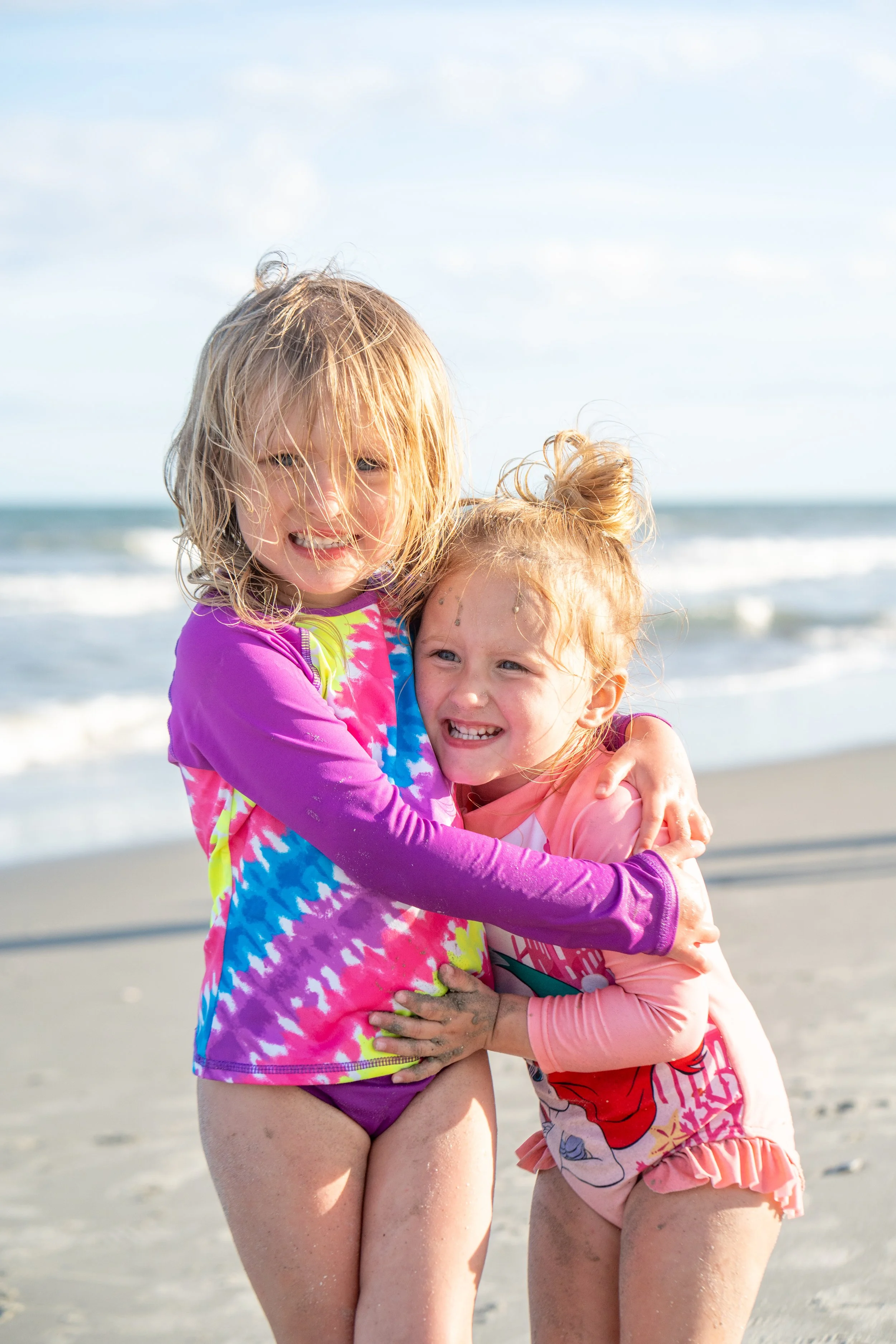 Two young girls hugging and smiling on the beach, wearing colorful swimsuits with the ocean and cloudy sky in the background.