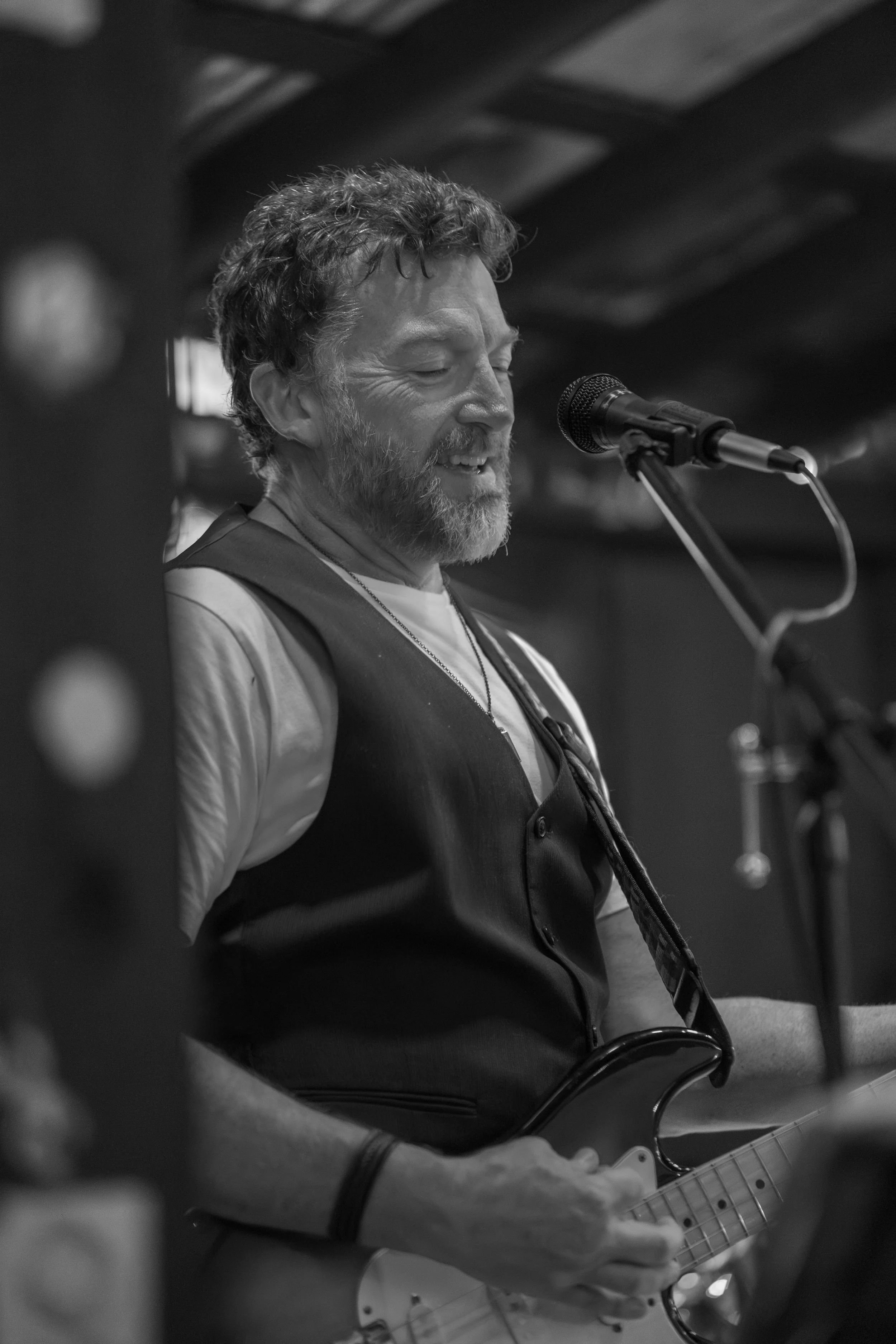 A black-and-white photo of a man with curly hair and a beard singing into a microphone while playing an electric guitar.