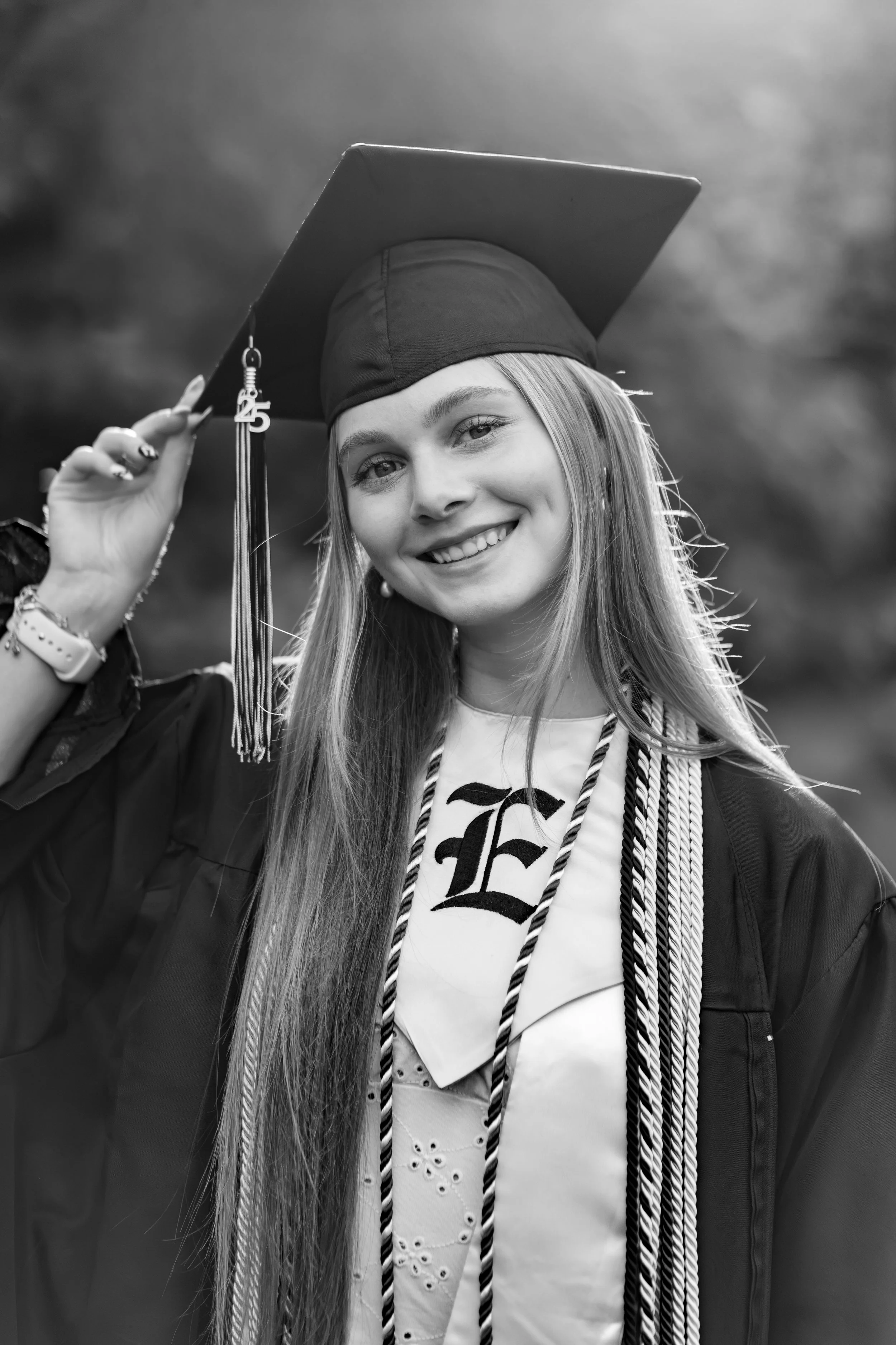 A young woman smiling, wearing a graduation cap and gown, with a tassel showing the number 25, and a decorated shirt underneath, outdoors.