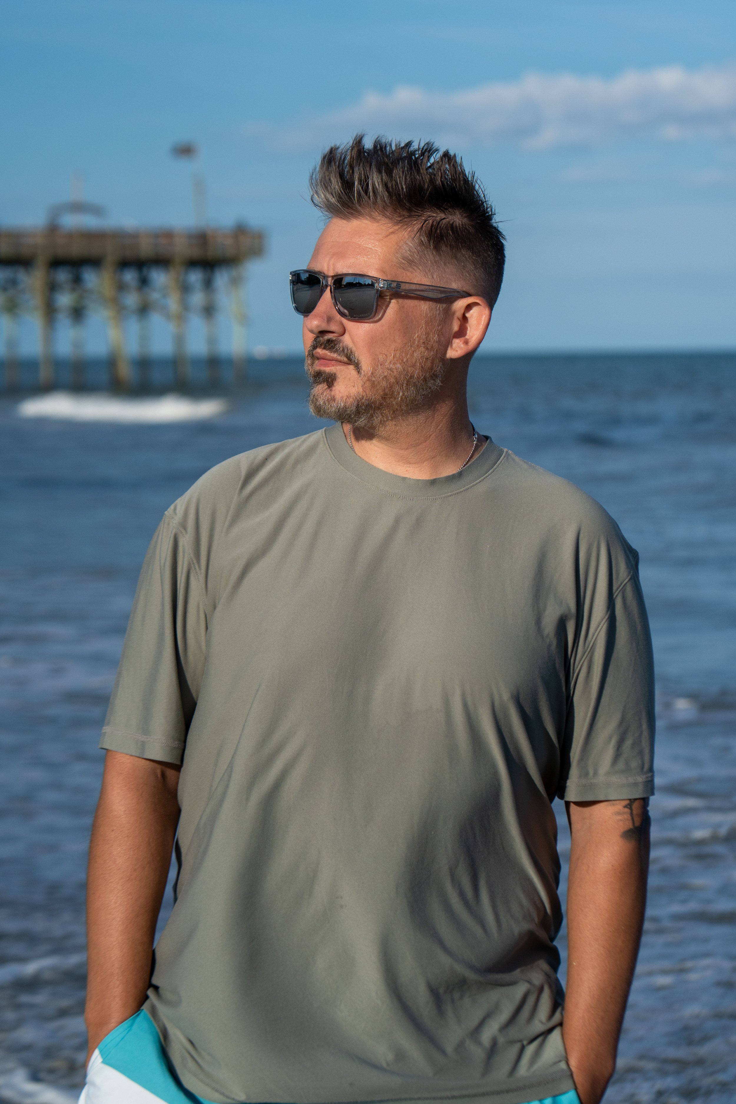 A man with sunglasses and a beard stands on the beach near the ocean with a pier in the background.