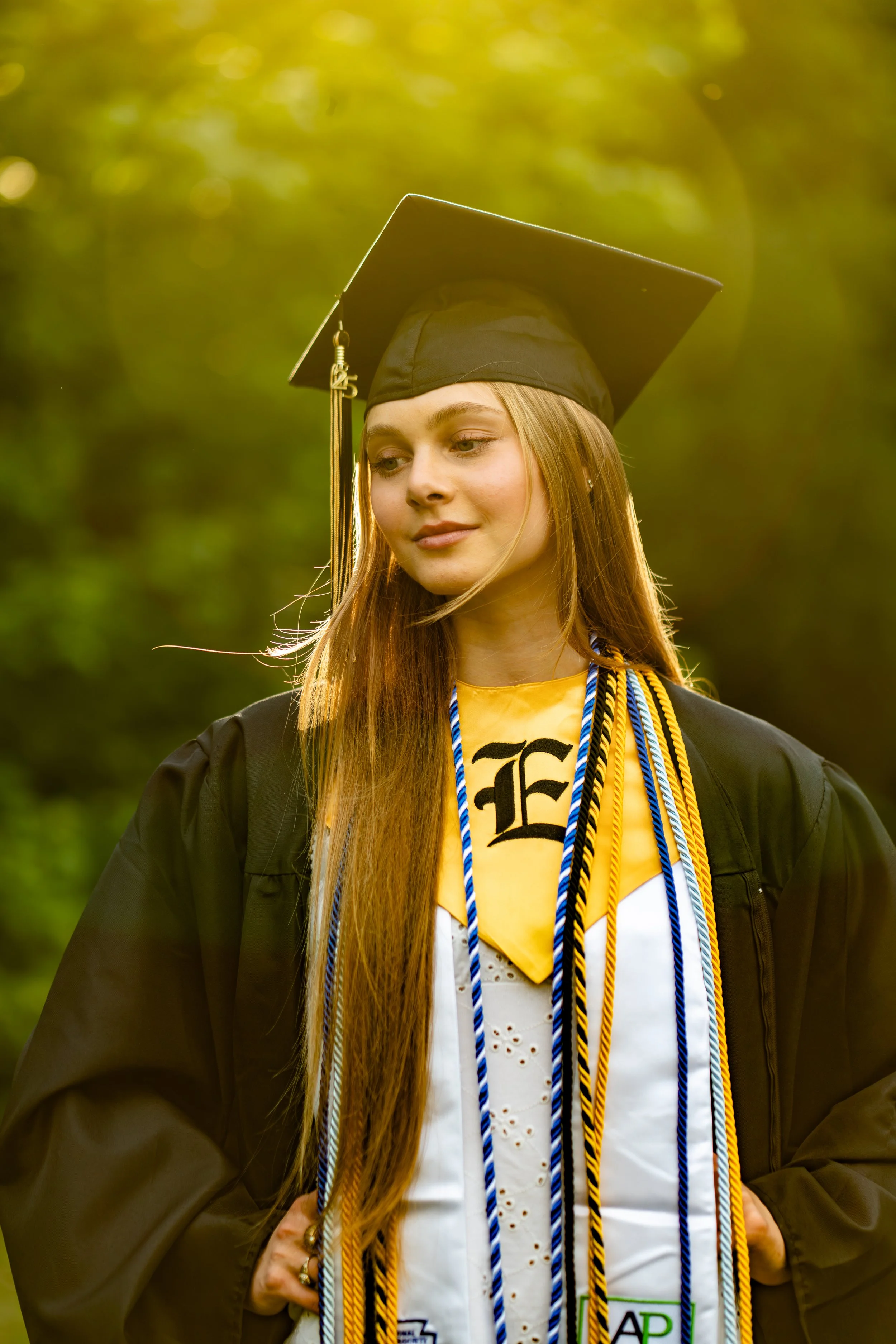 A young woman wearing a black graduation cap and gown, with multiple academic cords and medals, standing outdoors with blurred greenery in the background.