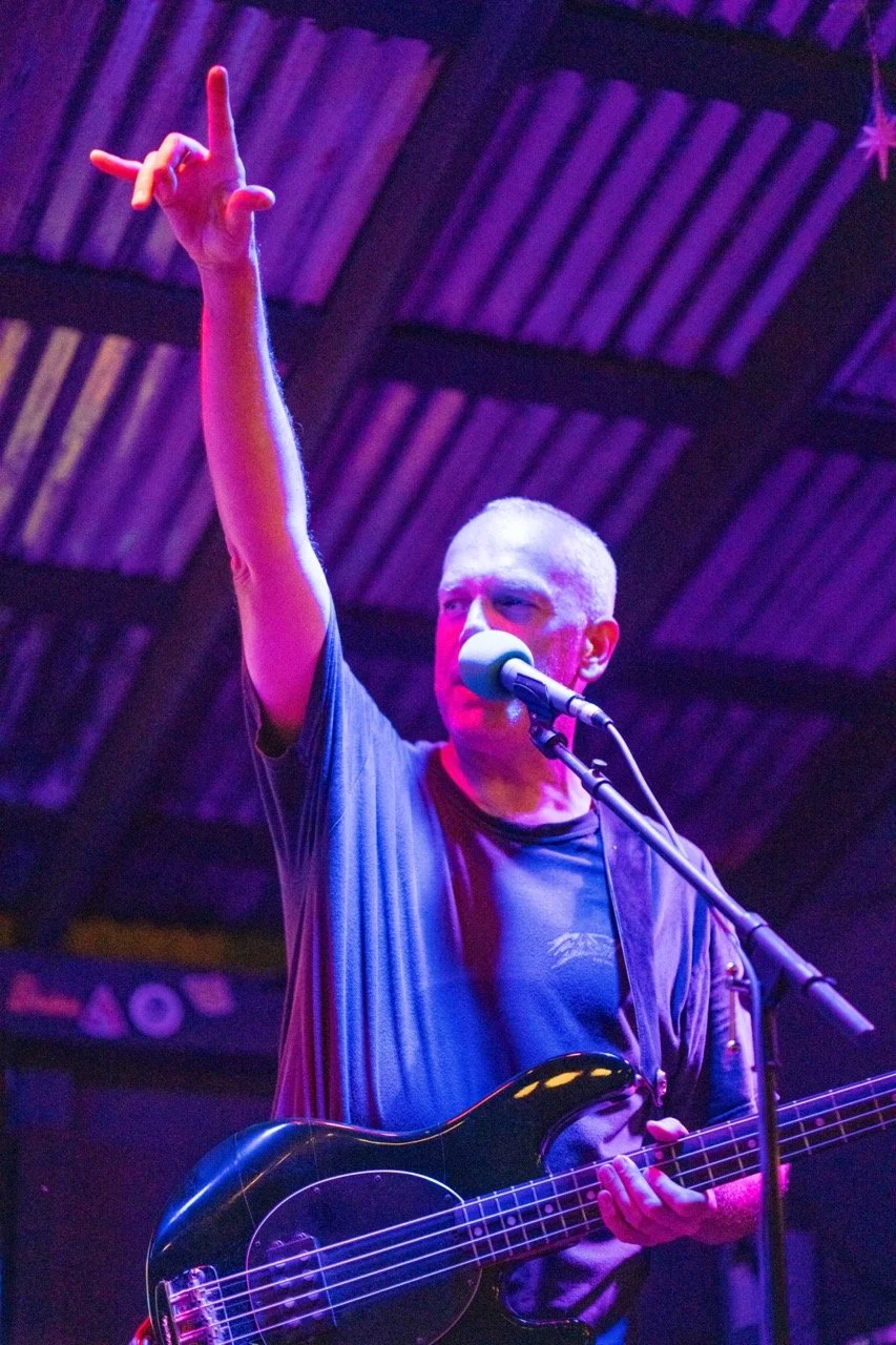 A man with short gray hair is on stage playing a black electric bass guitar and singing into a microphone, with his right hand raised in the air. The stage is lit with purple and pink lights, and a roof structure is visible in the background.