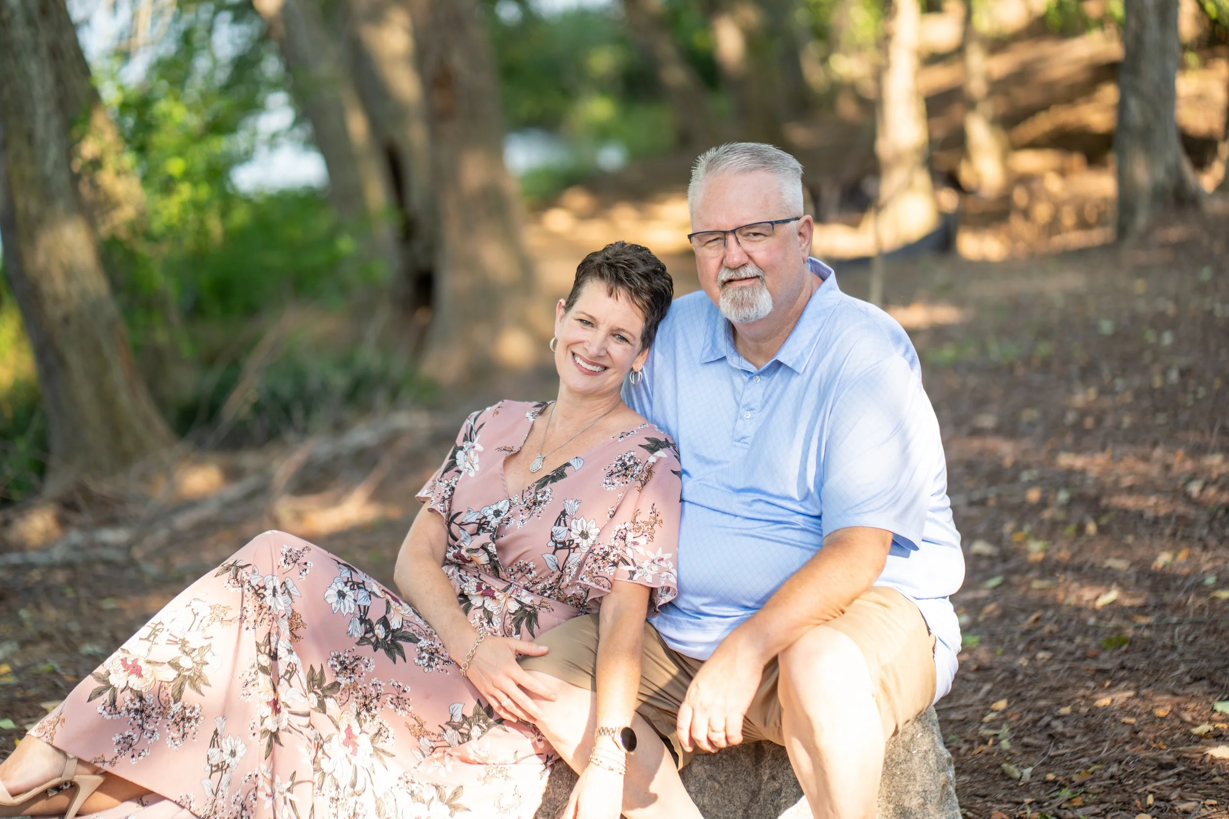A joyful middle-aged couple sitting close together outdoors in a wooded park, smiling at the camera. The woman is wearing a floral pink dress and the man is in a light blue shirt with glasses.