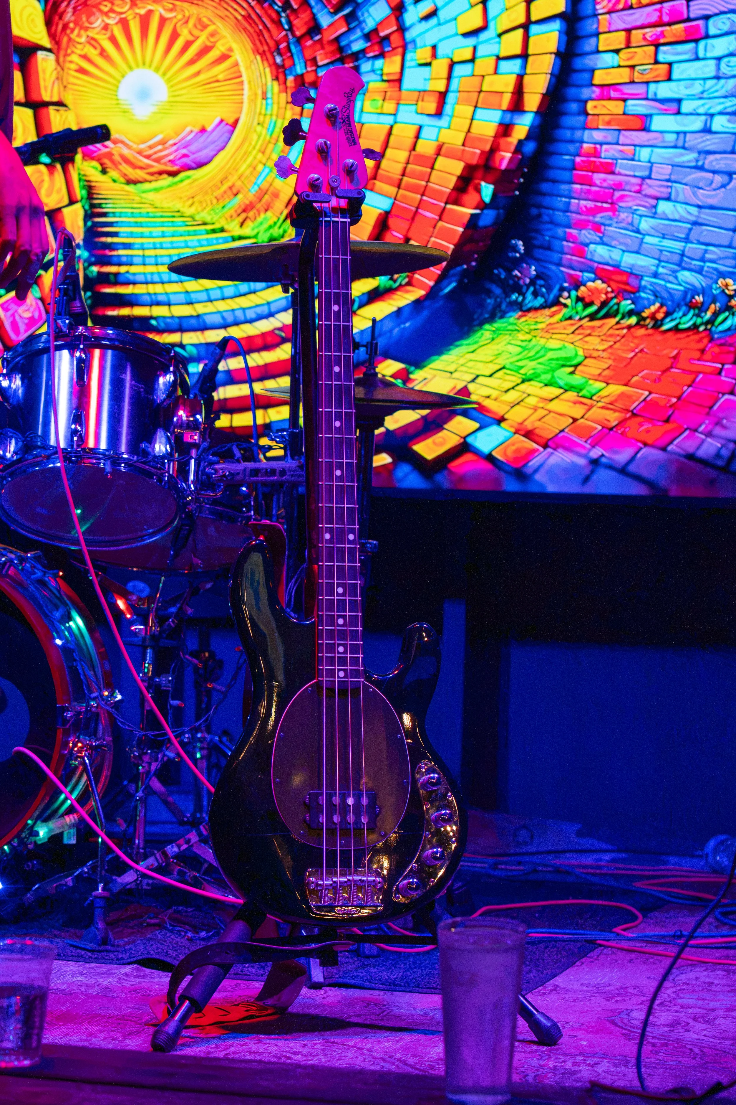 Stage with musical instruments including an electric bass guitar in the foreground and a drum set, illuminated by vibrant, colorful lighting with a digital backdrop displaying a psychedelic, swirling image of a sunrise over a landscape with brick-patterned hills and flowers.