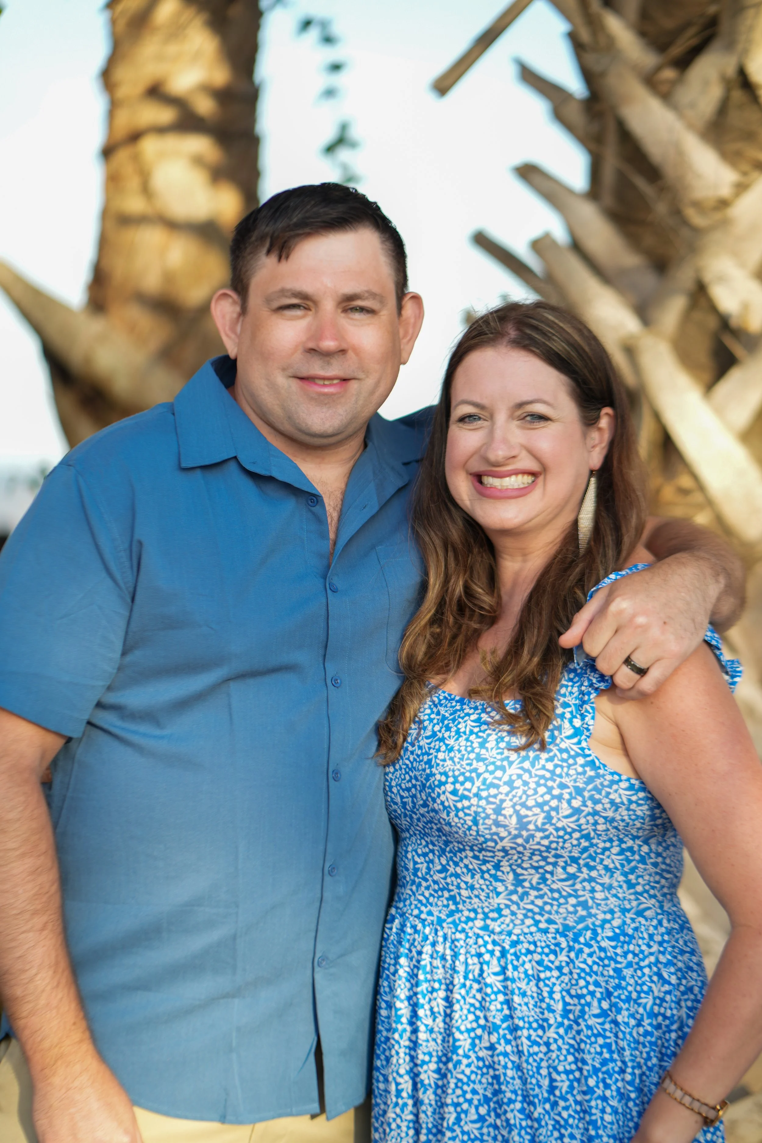 A smiling man and woman standing close together at an outdoor event, with a large tree with wooden structures in the background, both wearing casual summer clothing.