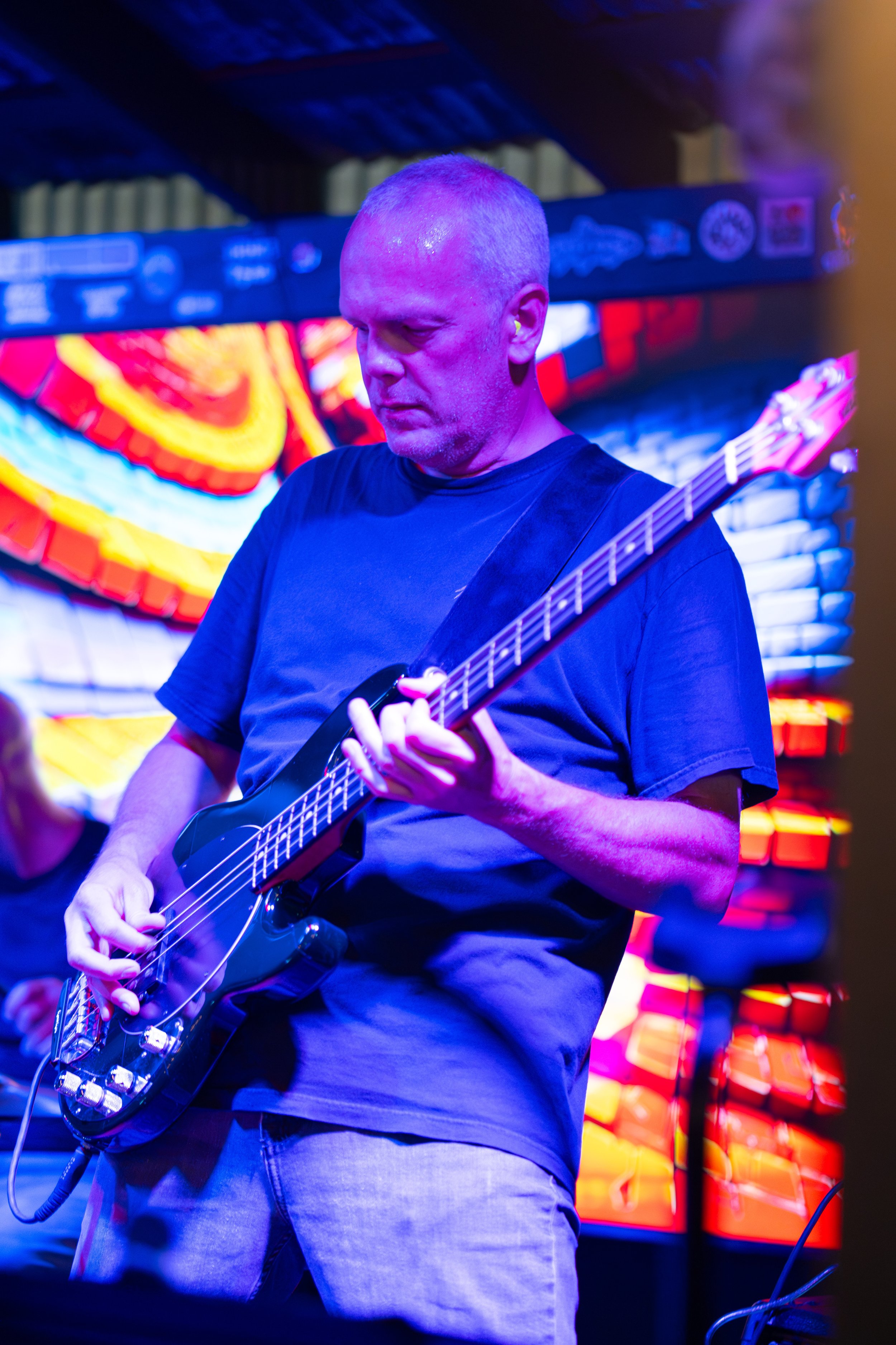 A man with a short haircut playing an electric guitar in a venue with colorful illuminated background.
