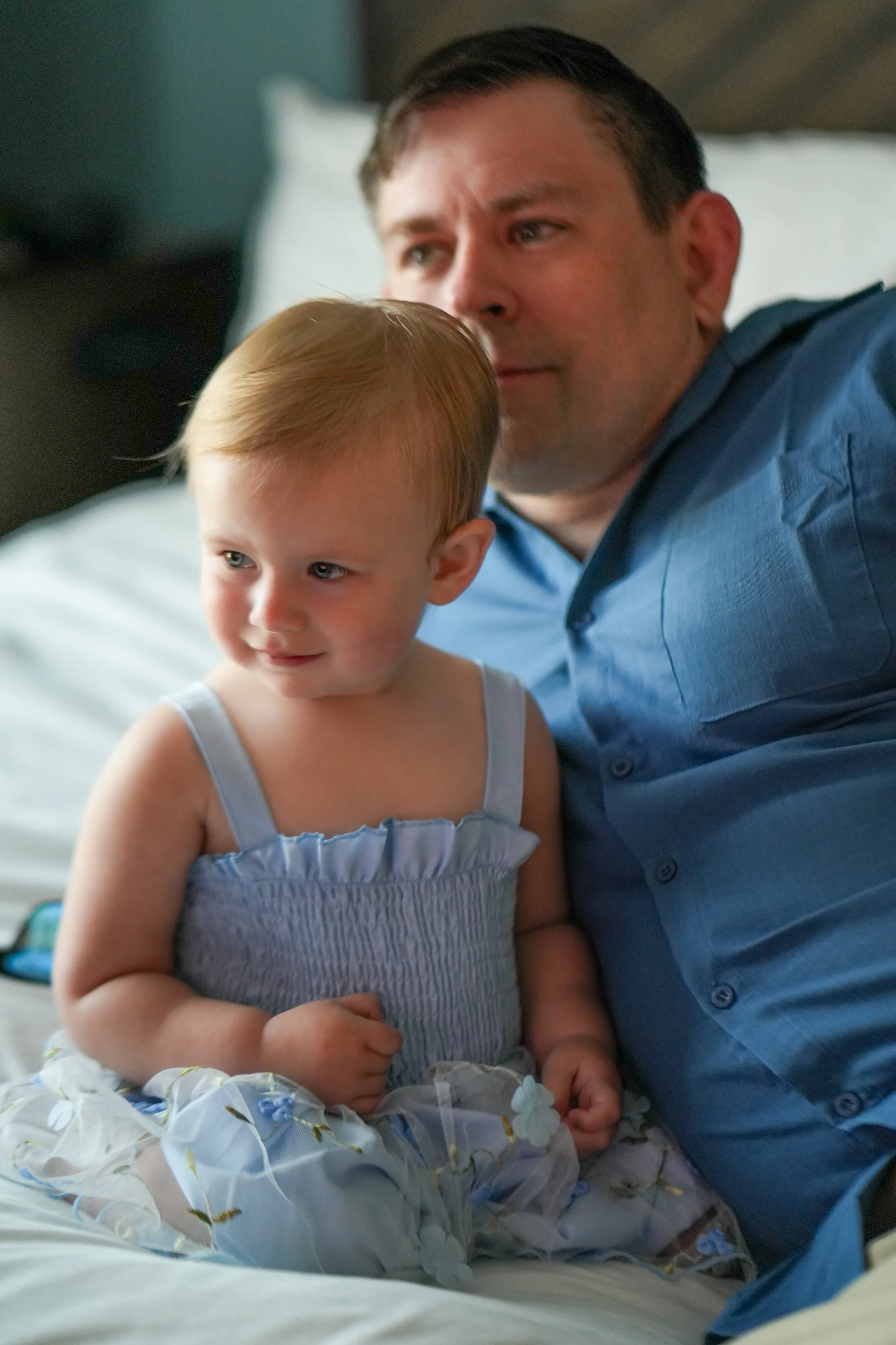 A man and a young girl sitting on a bed. The man is wearing a blue shirt, and the girl is wearing a light-colored dress with floral patterns. They are close together, with the man resting his face near the girl's head, in a relaxed, intimate moment.