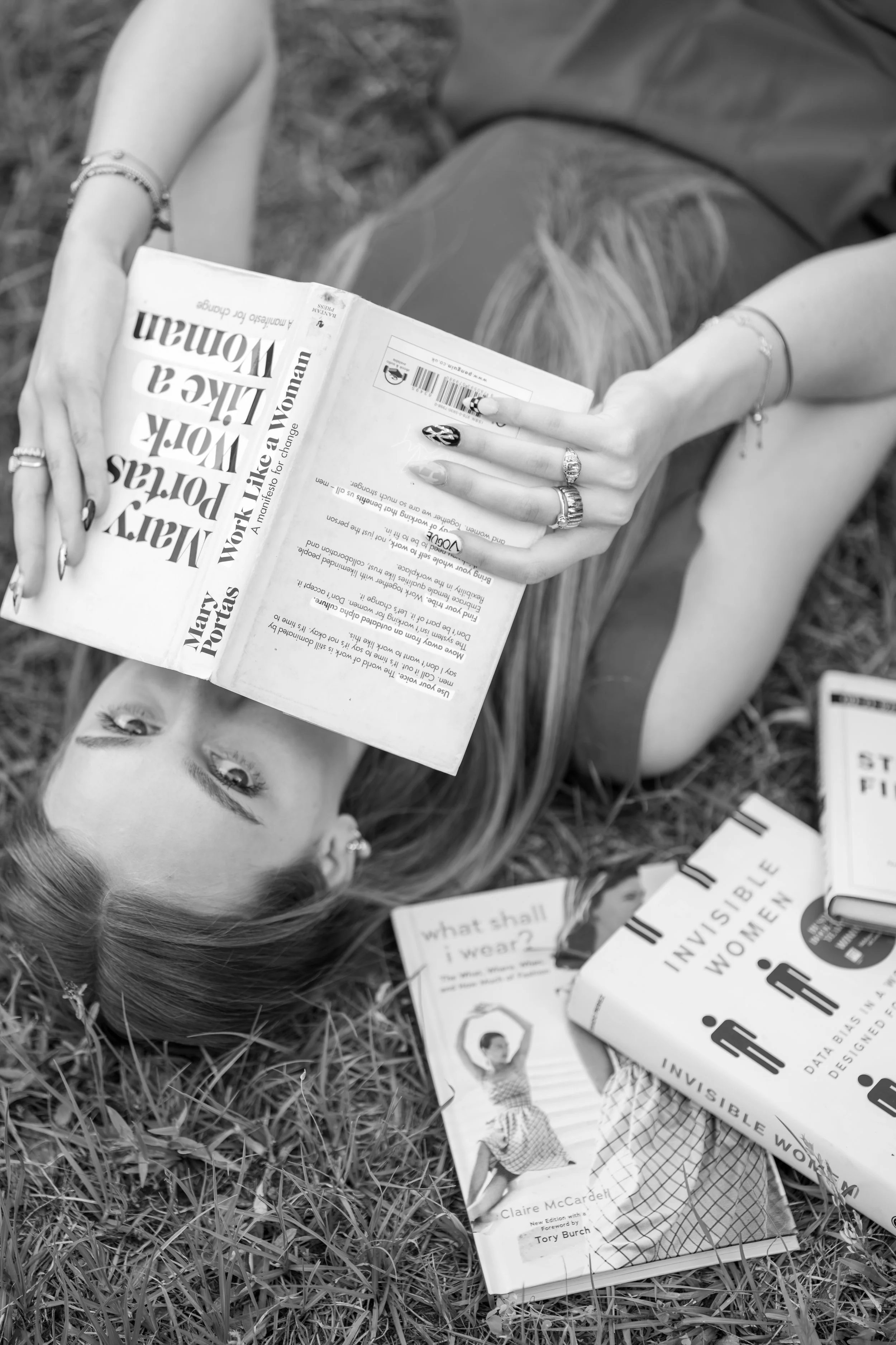 A person lying on the grass holding a magazine with messages about women and work, surrounded by additional magazines and papers on the ground.