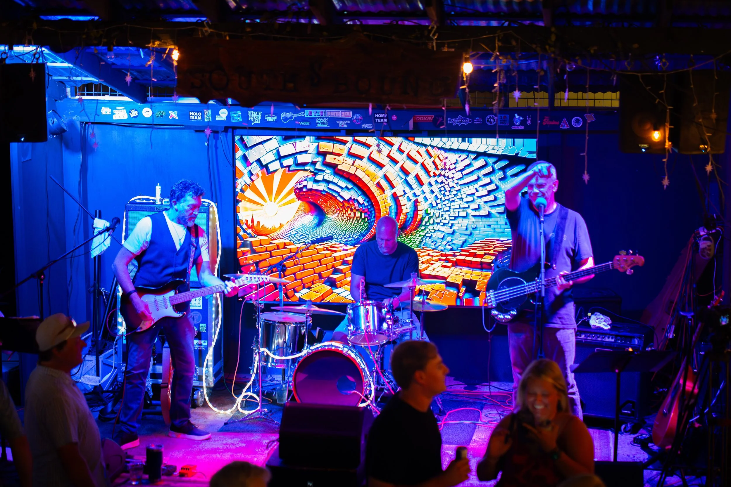 A band performing live on stage with a colorful digital backdrop. The band has three musicians, one playing guitar, one on drums, and another singing and playing bass guitar. Audience members are visible in the foreground.