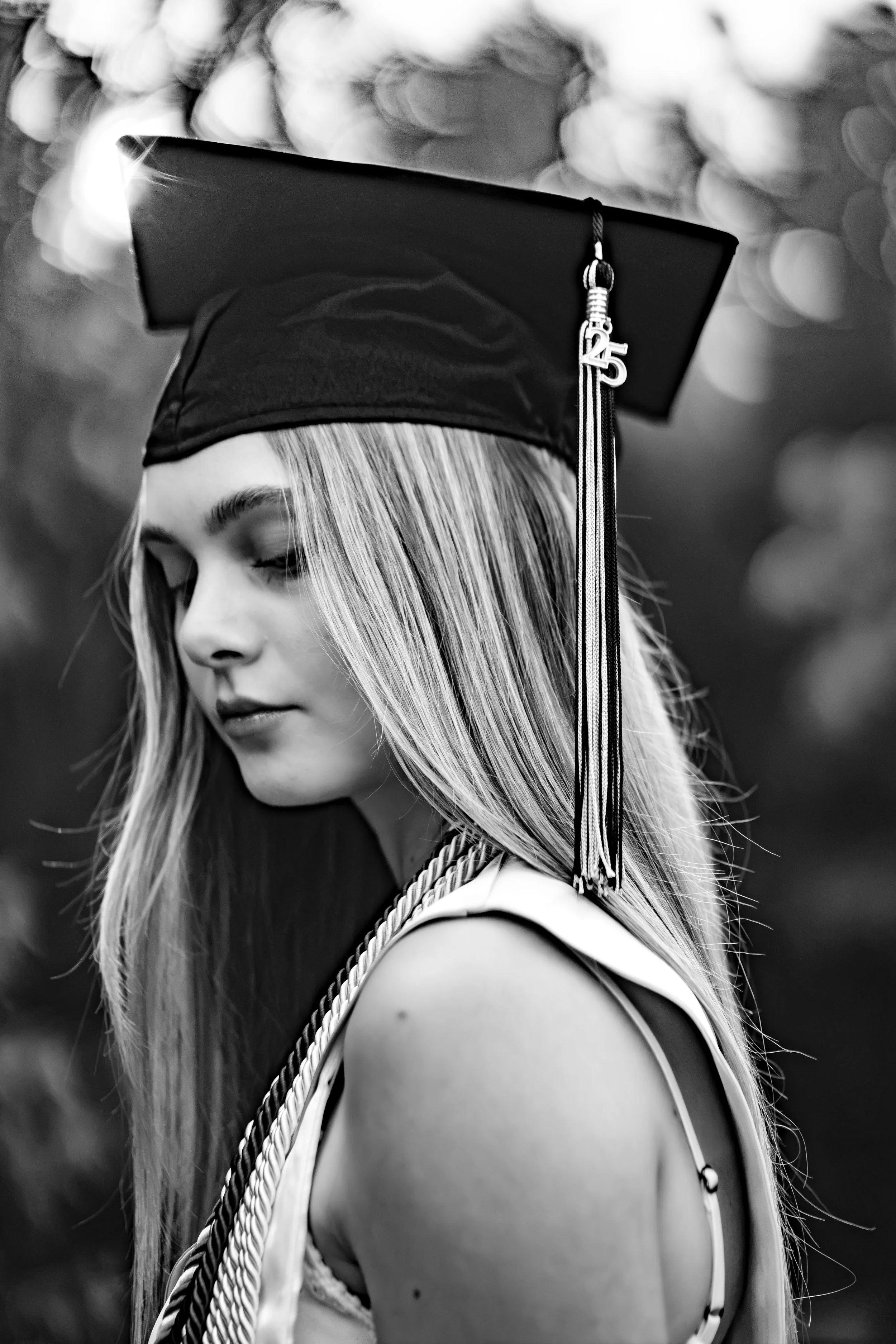 Black and white photo of a young woman wearing a graduation cap with a tassel that has a '25' charm, looking downward with long blonde hair, outdoors with blurred background.