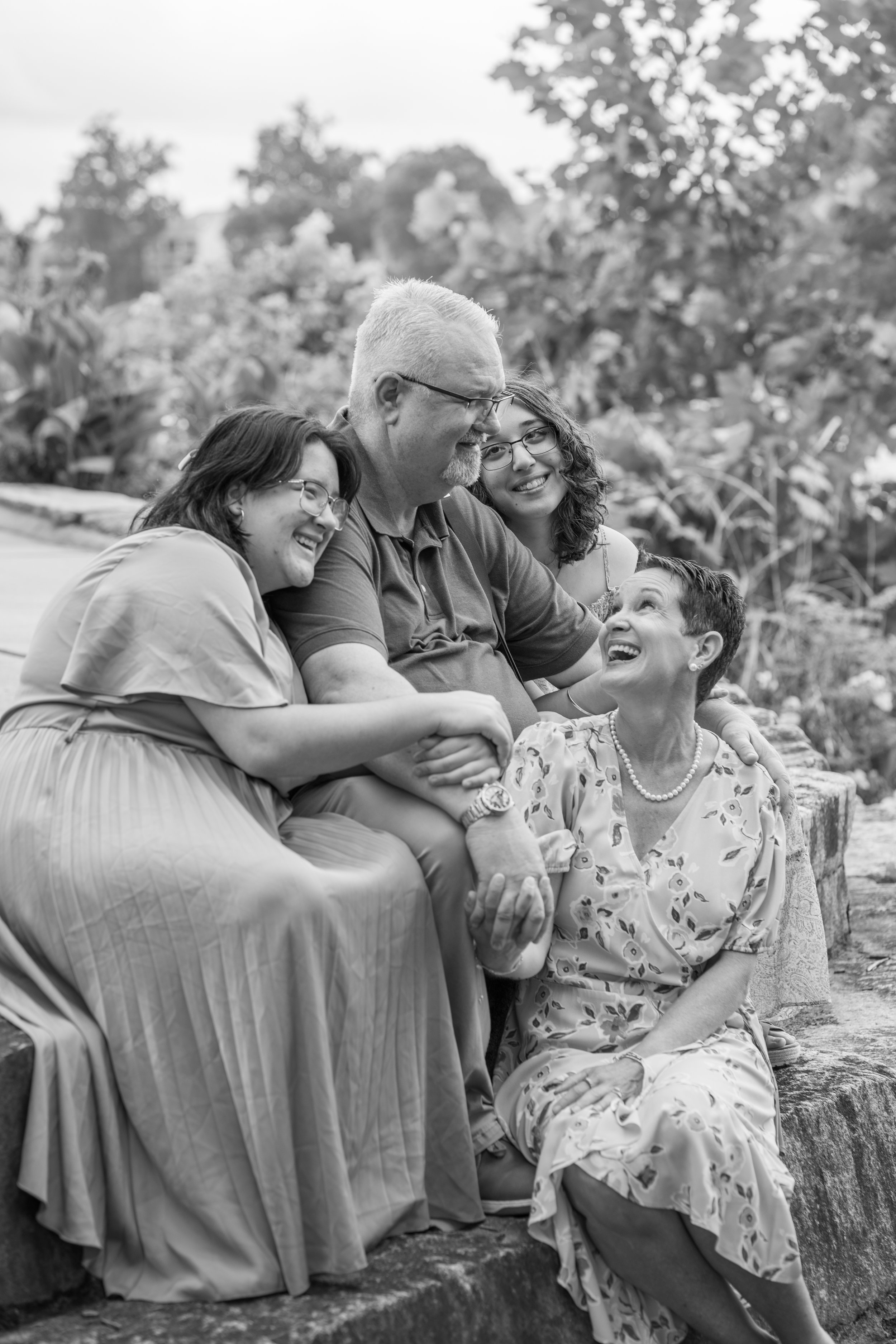 A group of five people, three women and two men, sitting close together outdoors, smiling and enjoying each other's company with a background of trees and foliage.