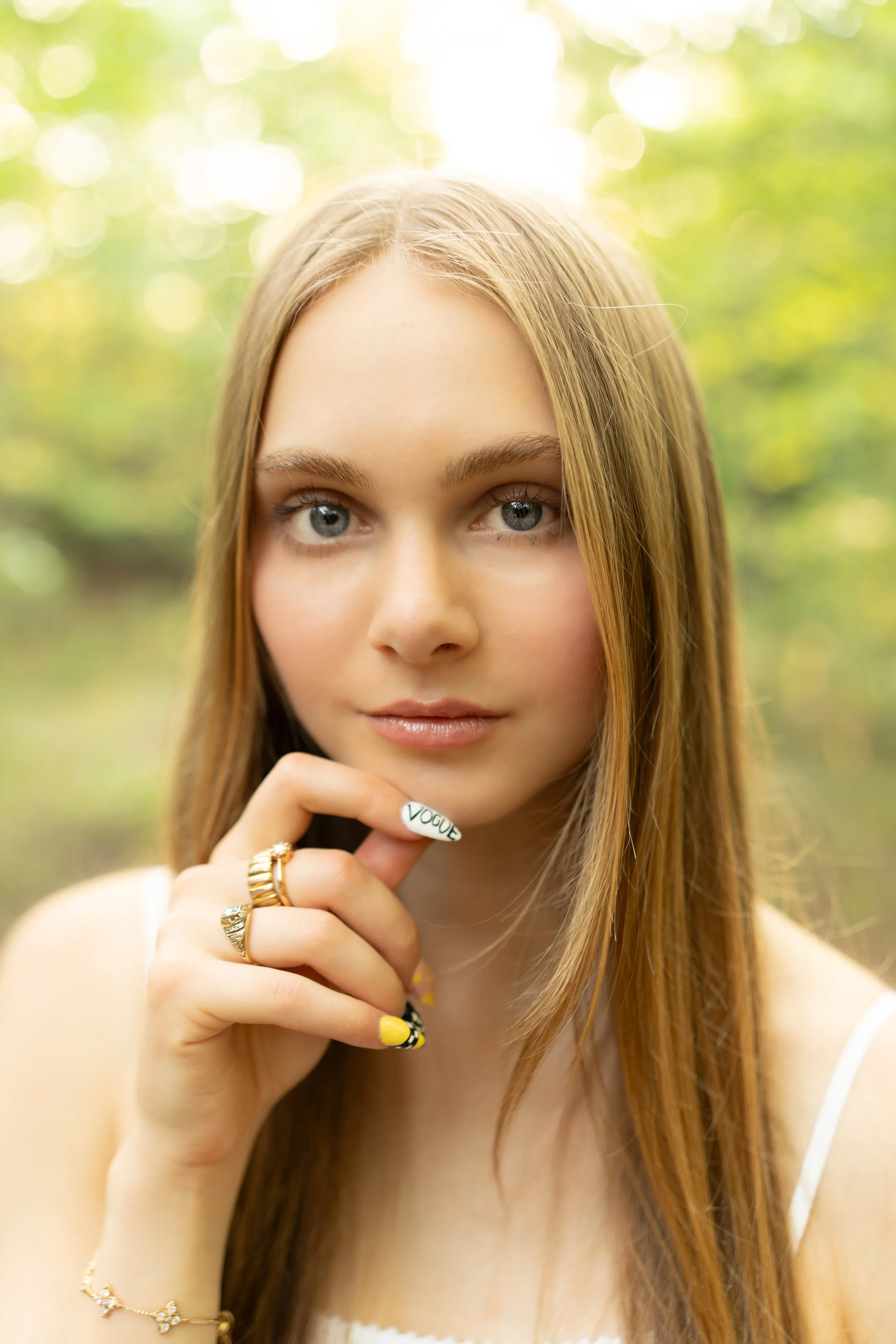 Close-up of a young woman with long, light brown hair and blue eyes, looking at the camera outdoors with blurred green trees and sunlight in the background.
