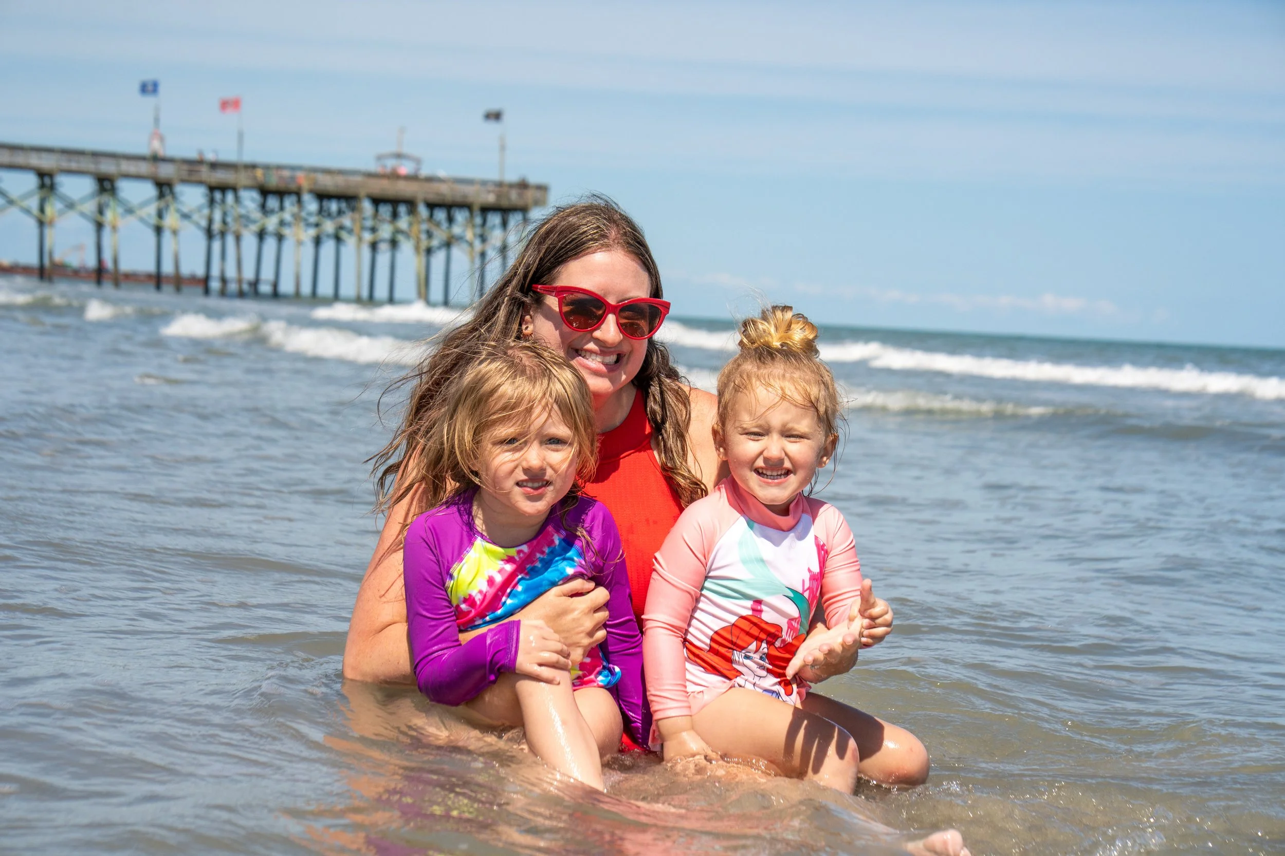 A woman with glasses and three young girls smiling and sitting in shallow ocean water near a pier on a sunny day.