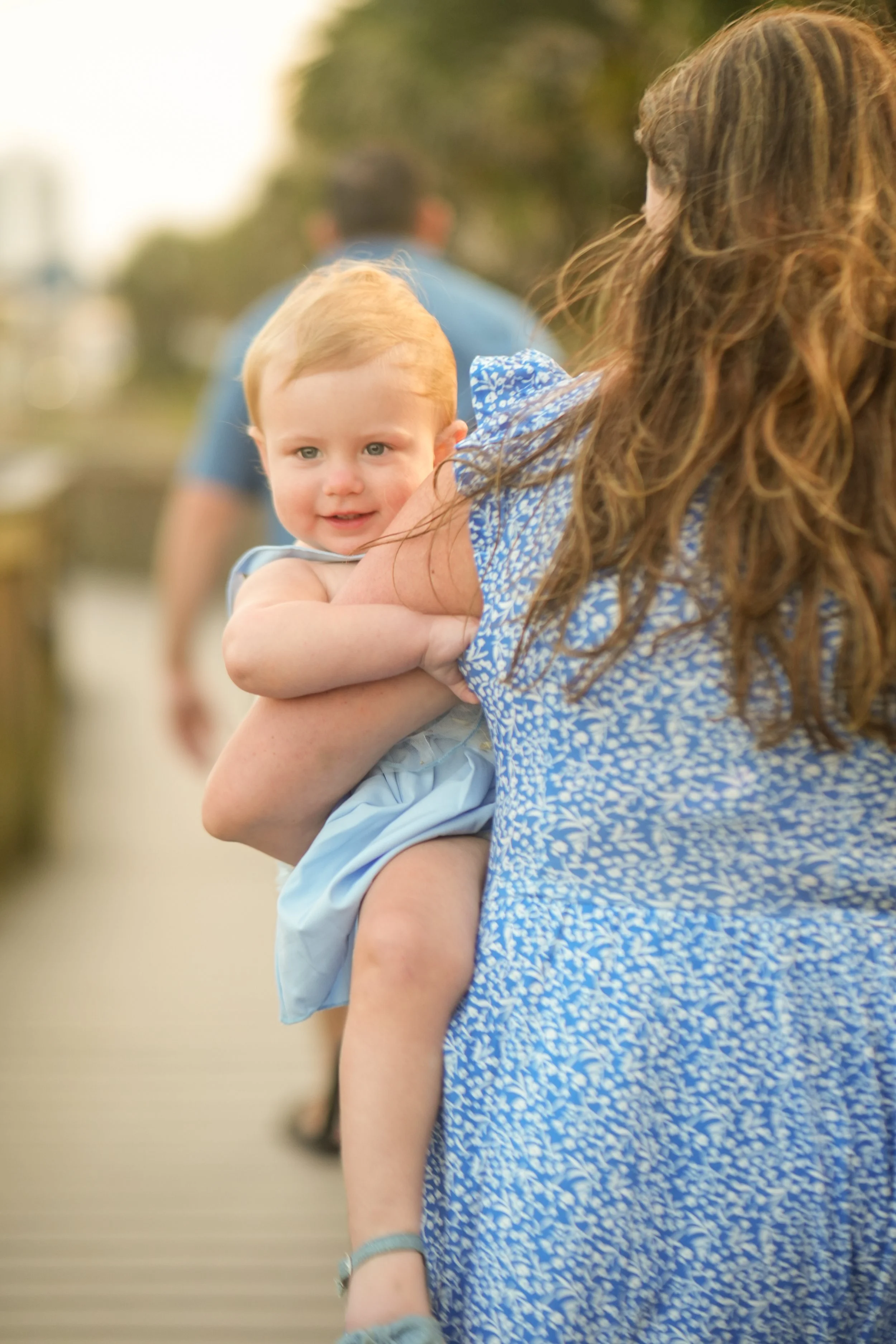 A woman with long, wavy brown hair is holding a young girl with red hair and blue eyes. The girl is smiling at the camera, wearing a blue floral dress. The background is blurred with people and outdoor scenery.