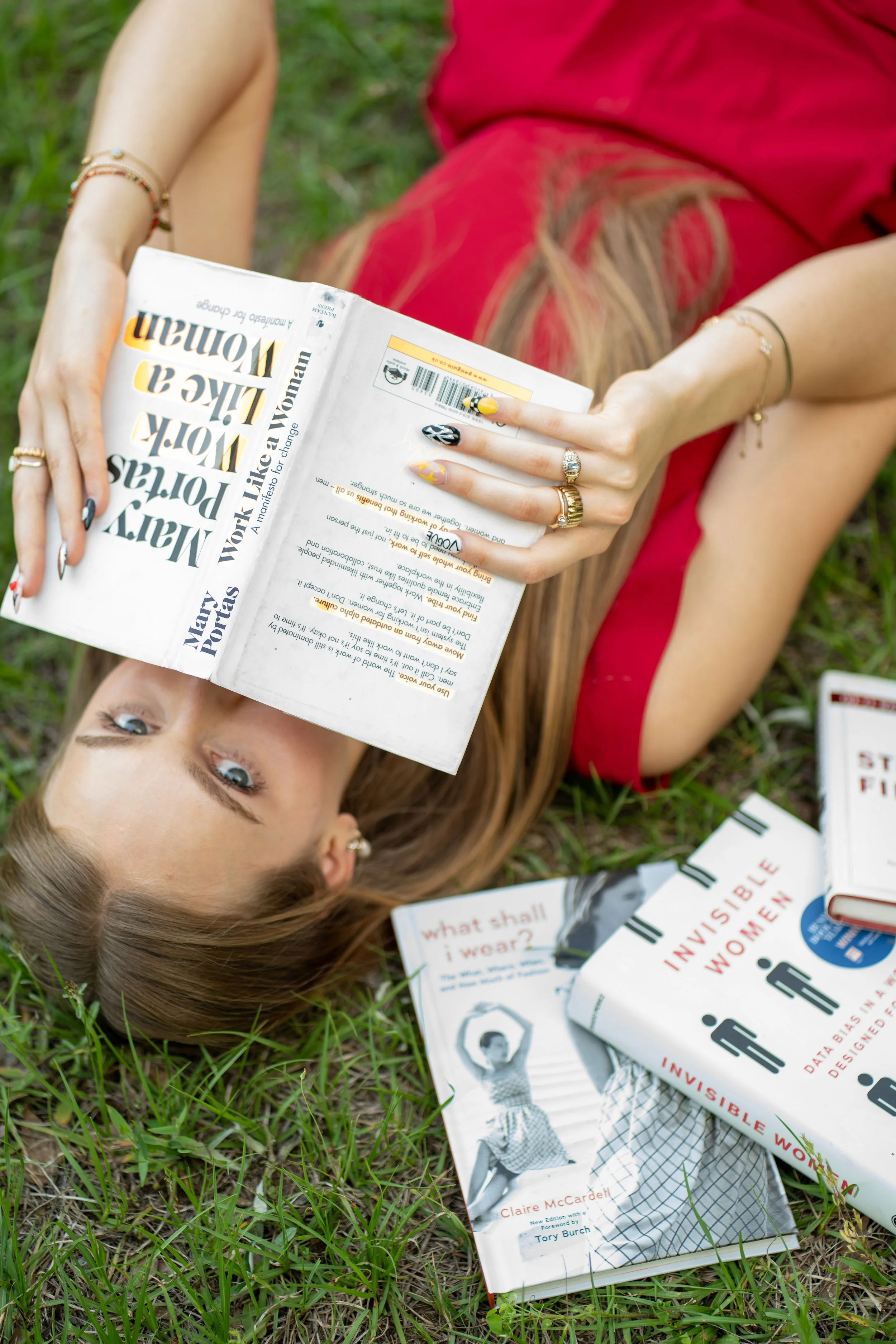 A woman with blonde hair lying on grass, holding a magazine titled "Work Like a Woman," with other books about women and invisible work scattered around her.