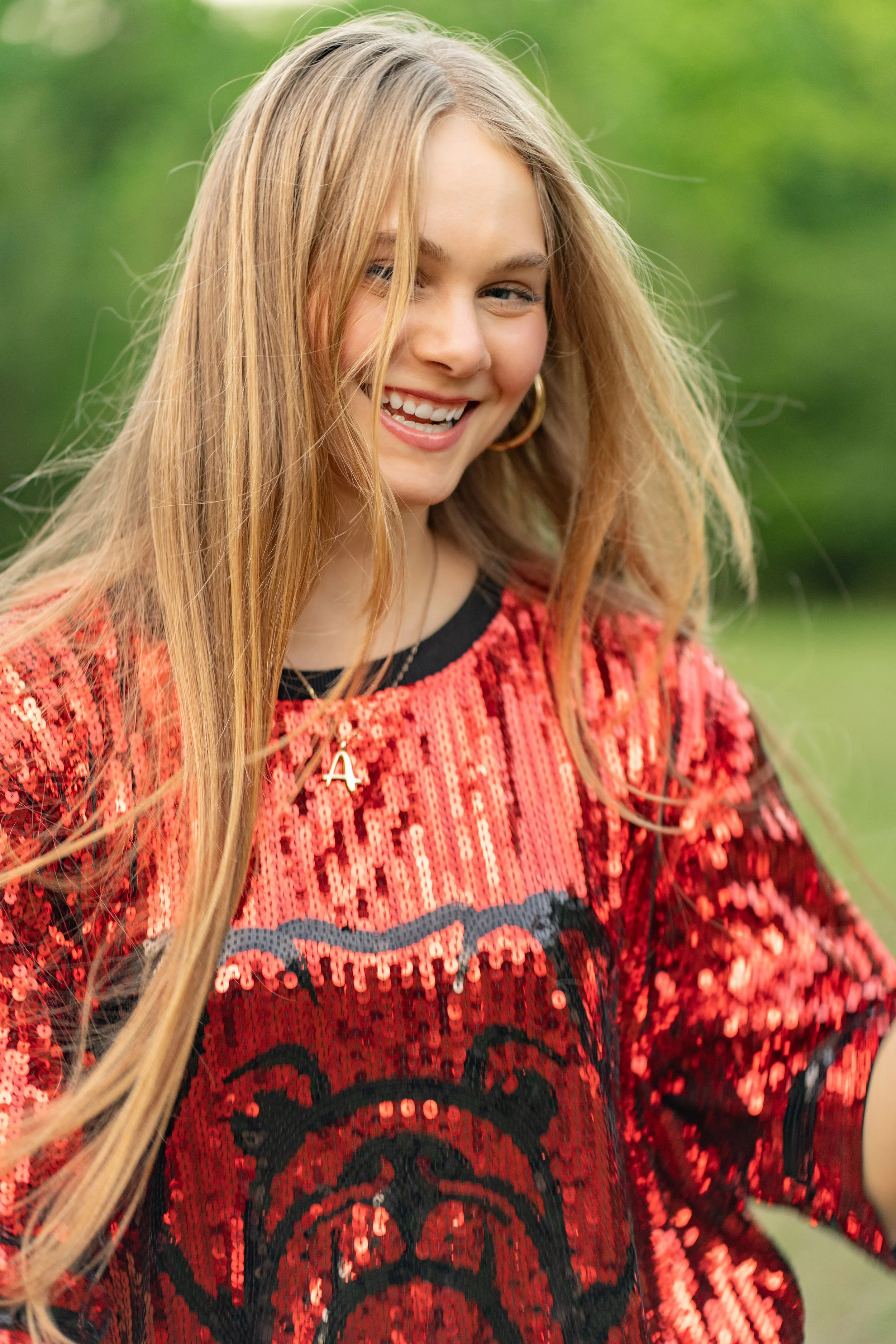 A young woman with long blonde hair, smiling and looking down, wearing a red and black sequined sweater with a letter 'A' necklace, in an outdoor setting with green blurred background.