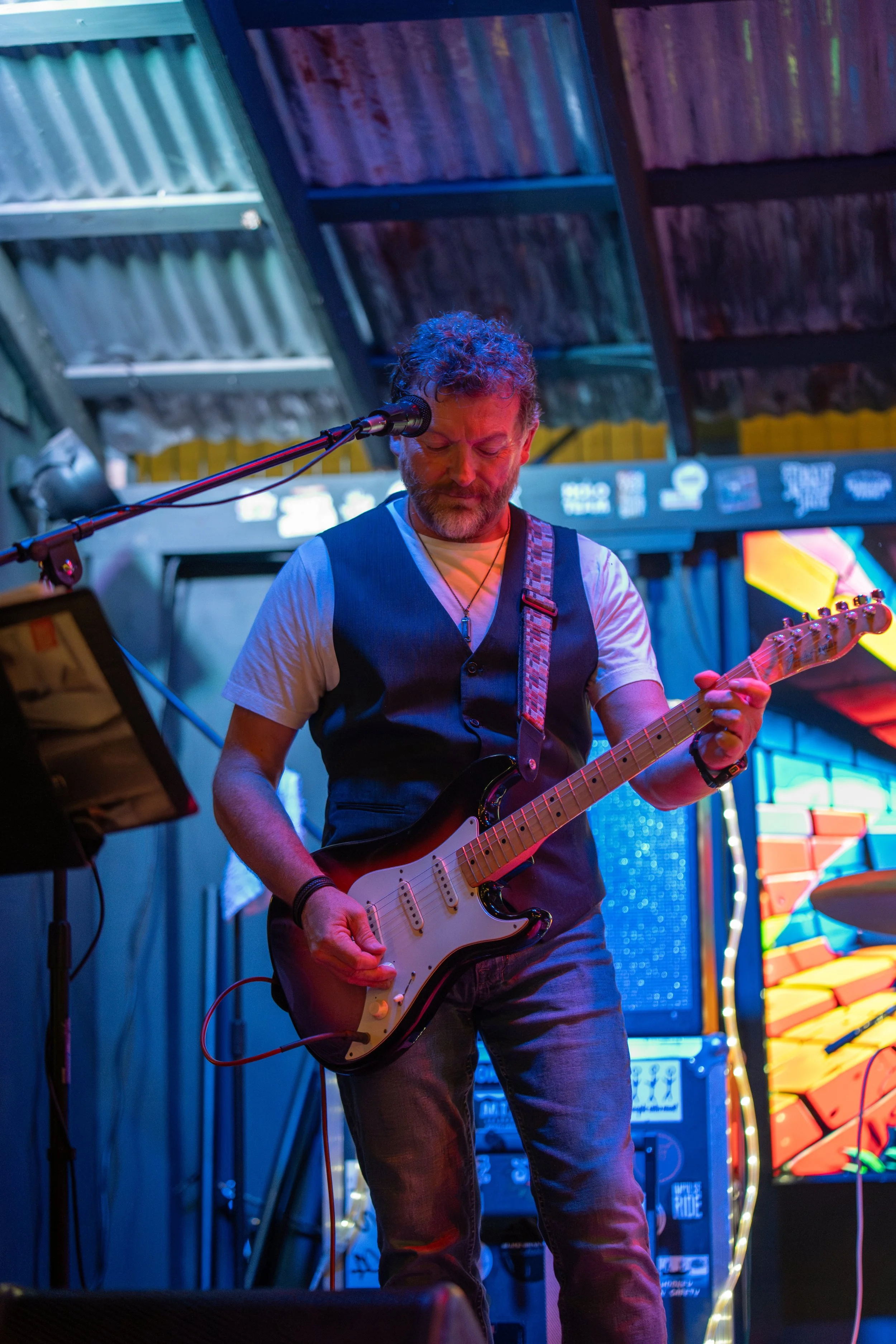 A man with curly hair and a beard playing an electric guitar on stage in a colorful, vibrant setting with neon lights and a music setup.