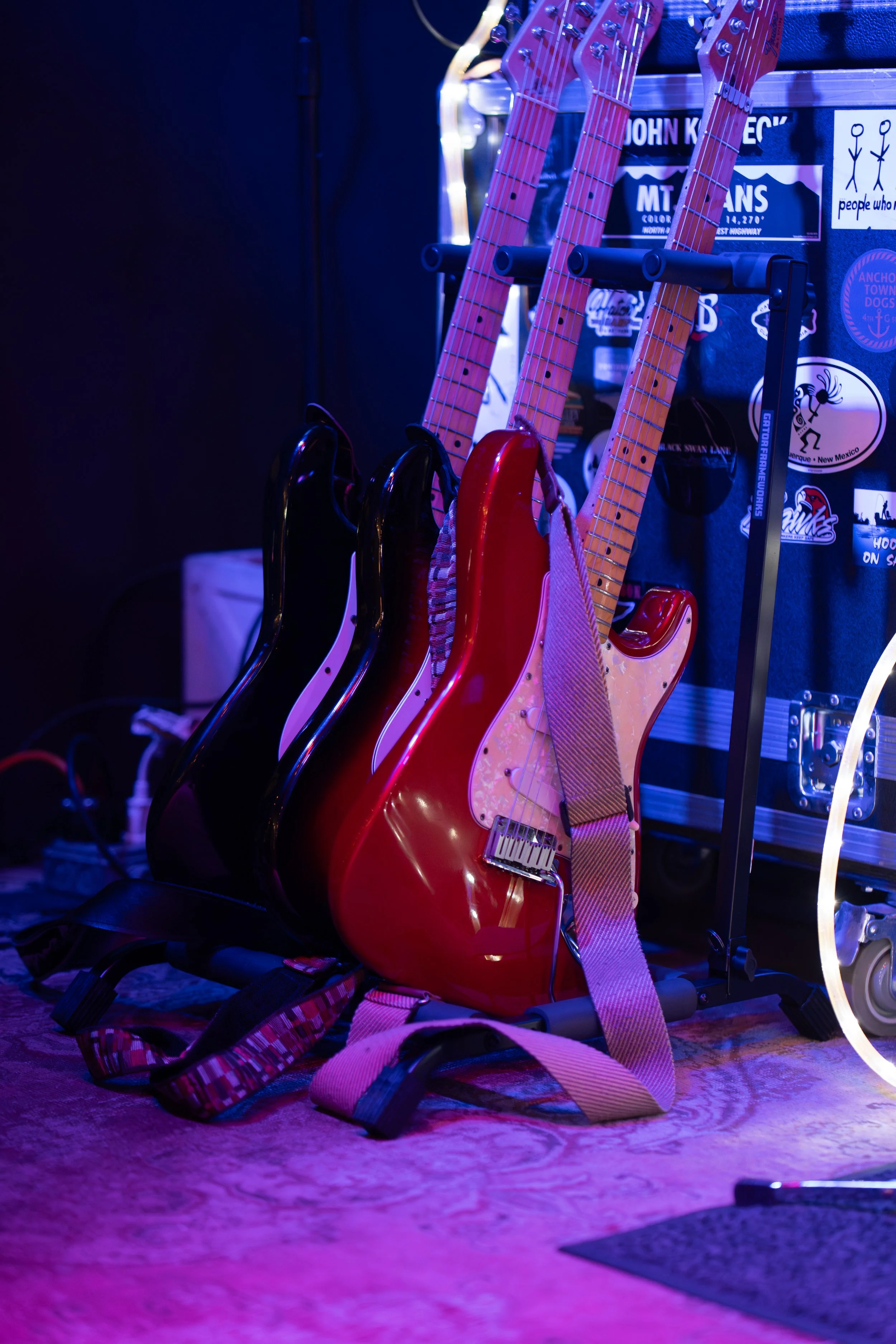 Four electric guitars with pink, red, and black finishes are on a stand, with a guitar strap draped over them, in a room illuminated by blue and purple lighting. There are stickers, a trailer, and other equipment in the background.