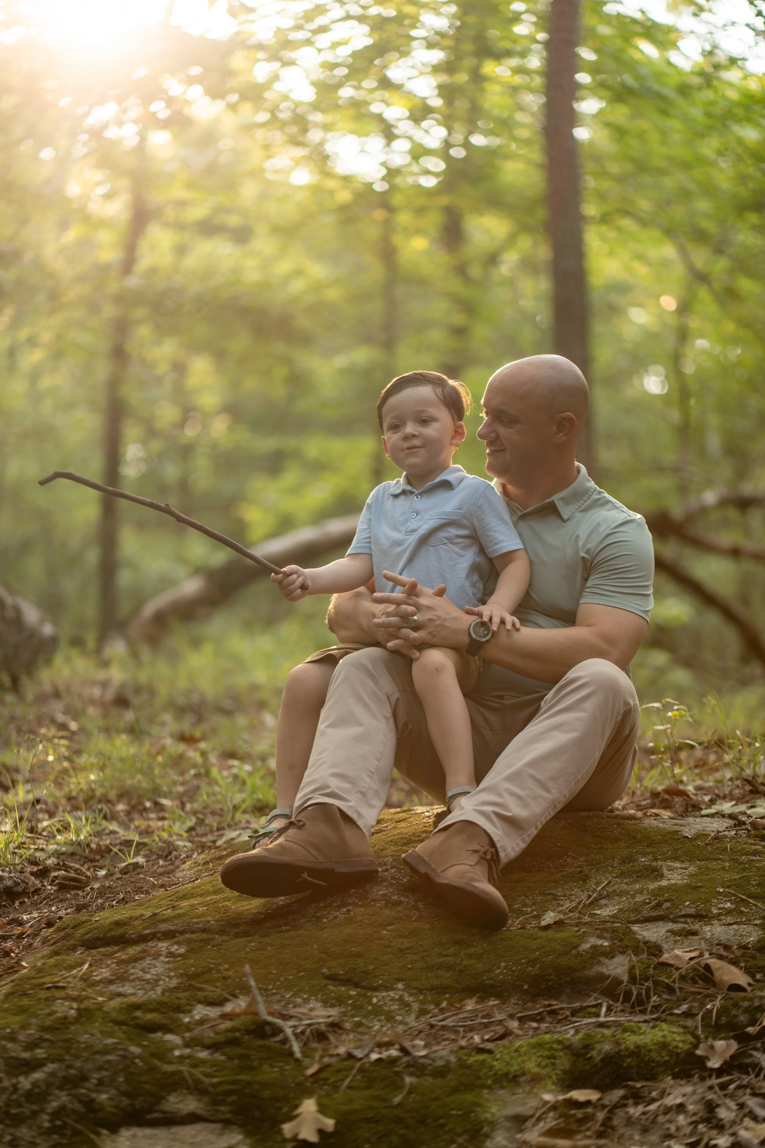 A man and a young boy sitting on a mossy log in a forest, with sunlight filtering through the trees, the boy is holding a stick.