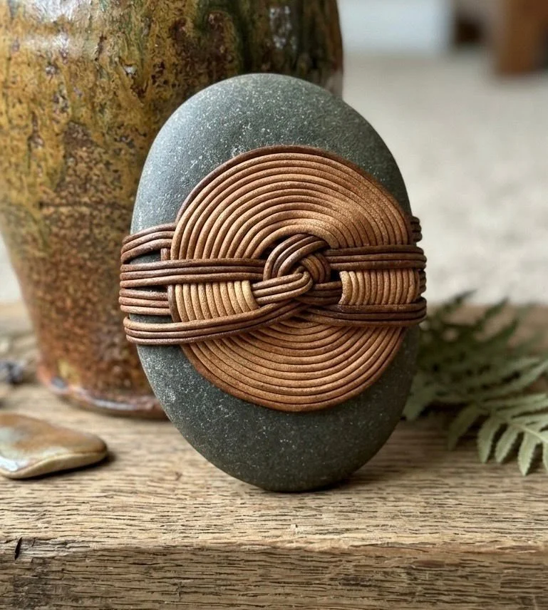 Decorative stone with intricately wrapped brown wire in a circular pattern, placed on a wooden surface with a tree trunk in the background.