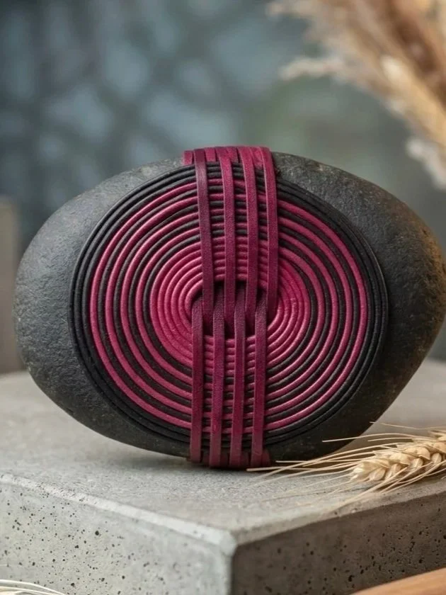 A round black yoga or meditation stone with a pink spiral pattern and five vertical pink bands wrapped around it, placed on a gray surface with some dried grass or wheat at the bottom right corner.