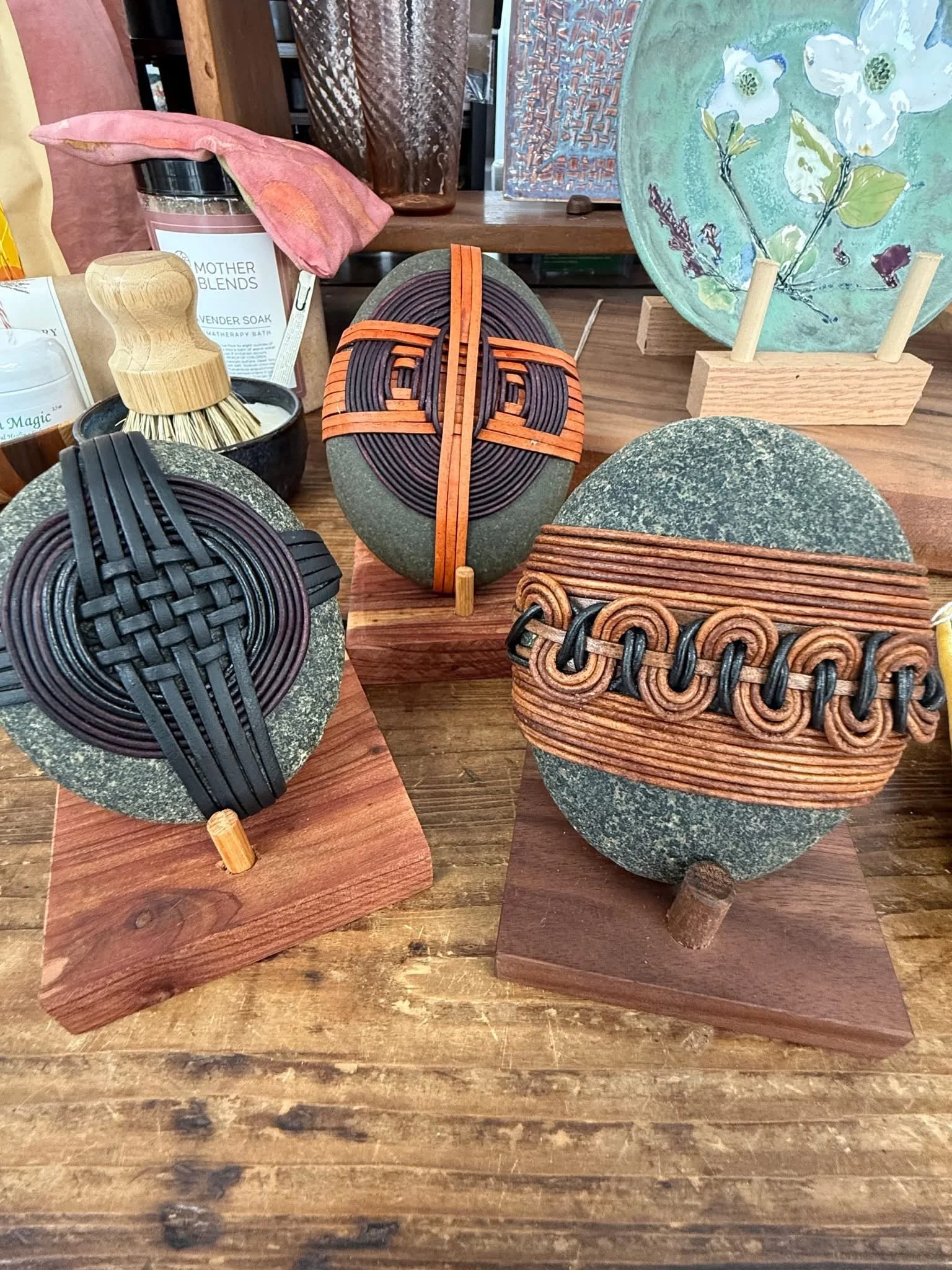 Decorative tabletop display with stones and woven objects, including a green stone with black and orange woven accents, and a small wooden stand holding a painted green plate with floral design, set on a wooden surface with a natural wood grain.