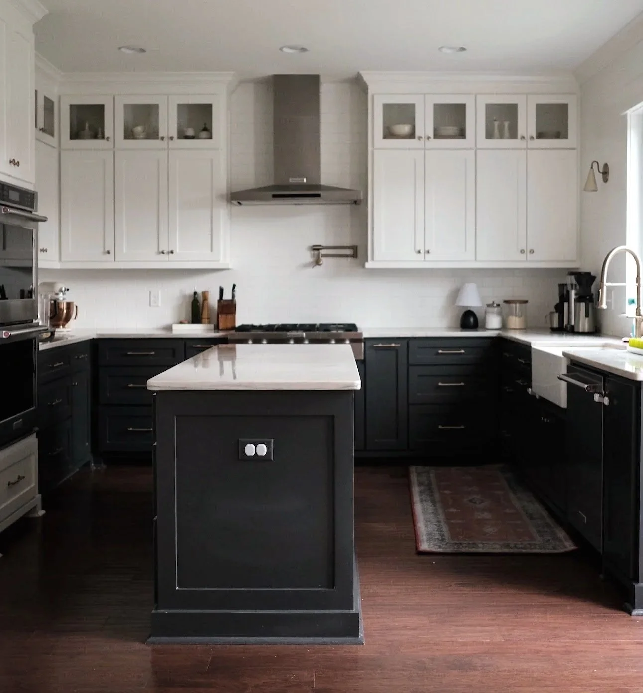 A modern kitchen with white upper cabinets and black lower cabinets, a white countertop, a kitchen island with a switch, and various appliances including a microwave, oven, and coffee maker. There are decorative items on the countertops and a patterned rug on the dark wood floor.