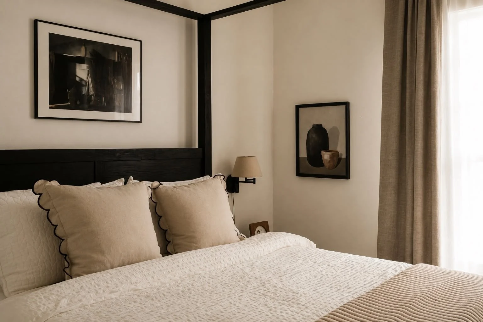 A neatly made bed with a quilted white cover and four beige pillows with dark trim in a bedroom. There is a wall-mounted lamp and framed artwork above the bed. A window with beige curtains is on the right.