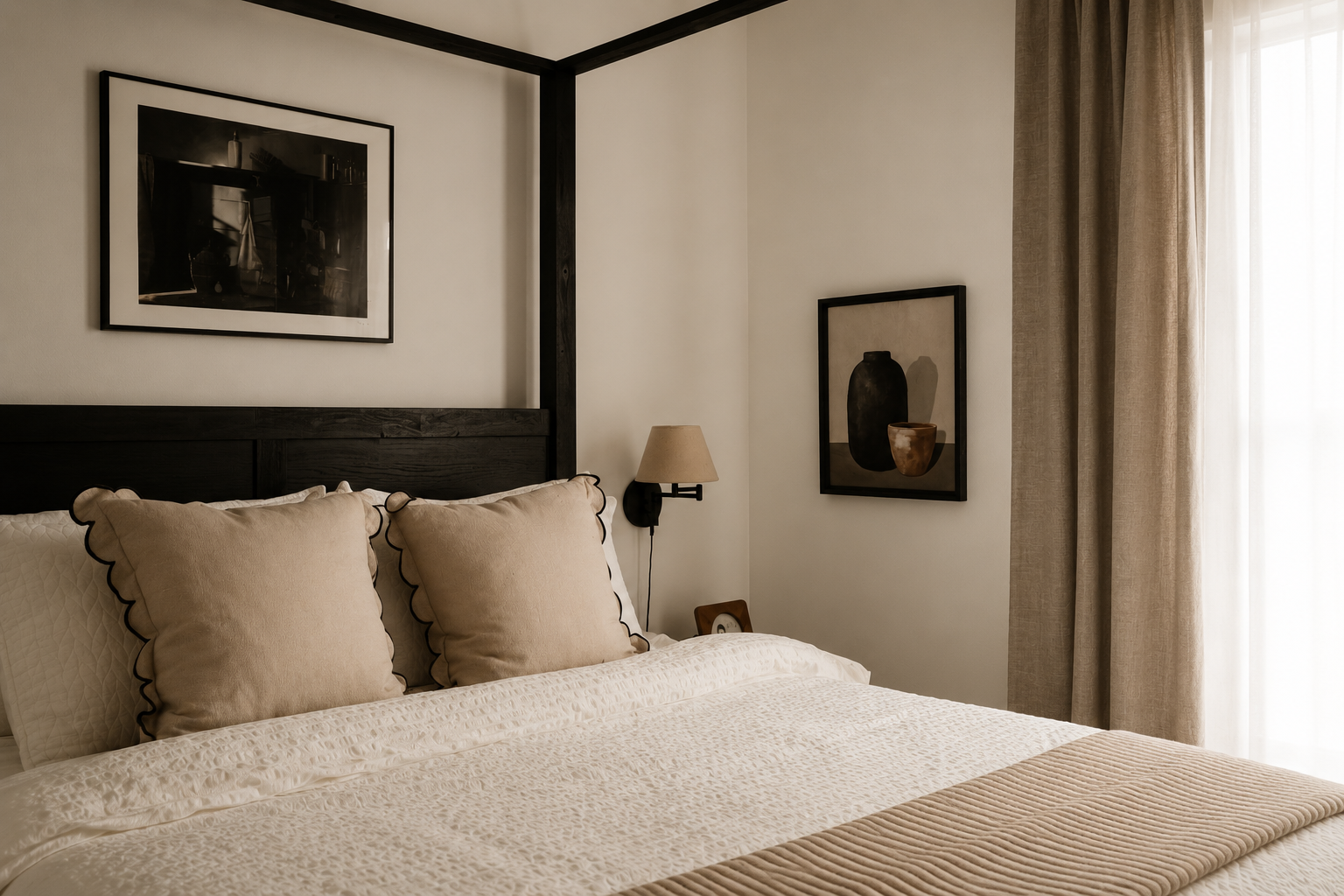 A neatly made bed with beige pillows and a beige blanket in a bedroom with framed black-and-white art on the walls and a window with beige curtains.