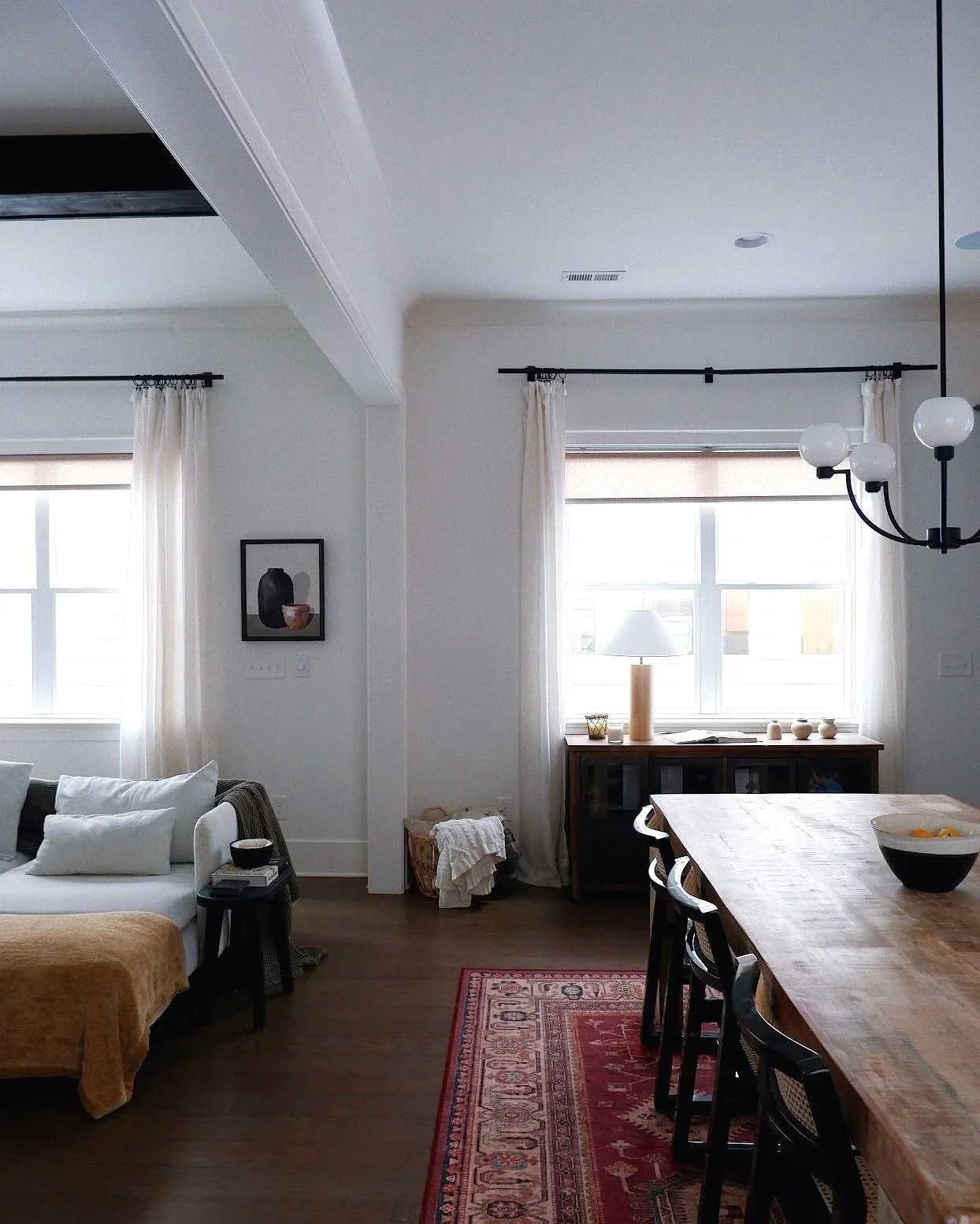 Living room and dining area with white walls, large windows with sheer curtains, a wooden dining table with black chairs, a sofa with white pillows, a sideboard with decor, a lamp, and ceiling light fixtures.