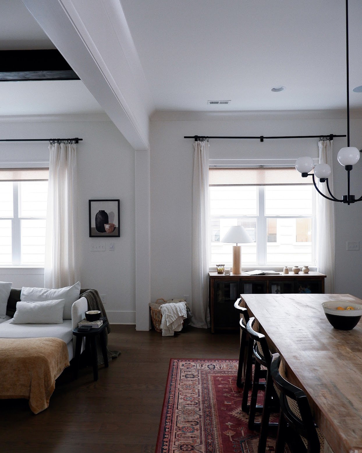Living room and dining area with white walls, large windows with sheer curtains, a wooden dining table with black chairs, a sofa with white pillows, a sideboard with decor, a lamp, and ceiling light fixtures.