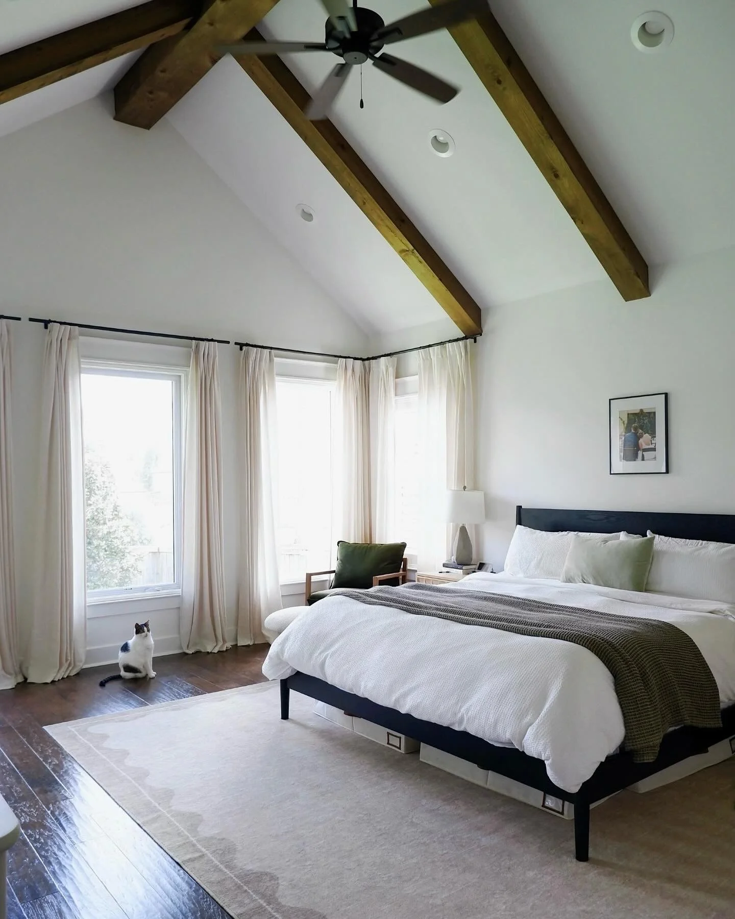 A bedroom with a vaulted ceiling featuring wooden beams and a ceiling fan. There are large windows with cream-colored curtains, and a black and white cat sitting on the floor near the window. The bed has white bedding, a brown blanket, and several pillows. A side table with a lamp and framed photograph is next to the bed.