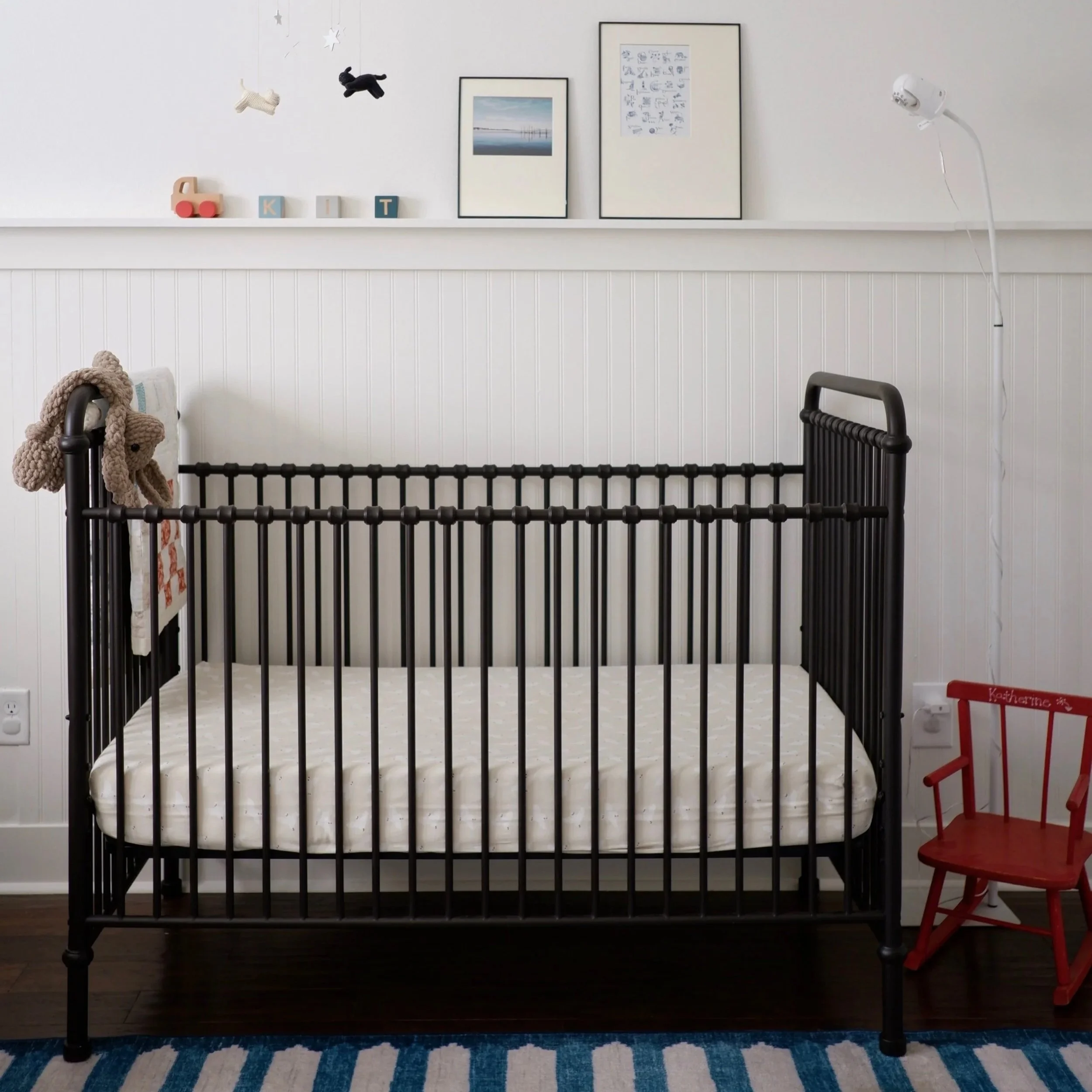 A black metal crib with a white mattress in a room with white walls and dark hardwood flooring. To the right of the crib is a red wooden chair and to the left, a beige plush teddy bear is hanging on the crib's side. A blue and white striped rug is under the crib. On the wall behind the crib, there is a white shelf with decorative framed pictures, small stuffed animals hanging from the ceiling, and colorful letter blocks spelling parts of the name 'KIT'.