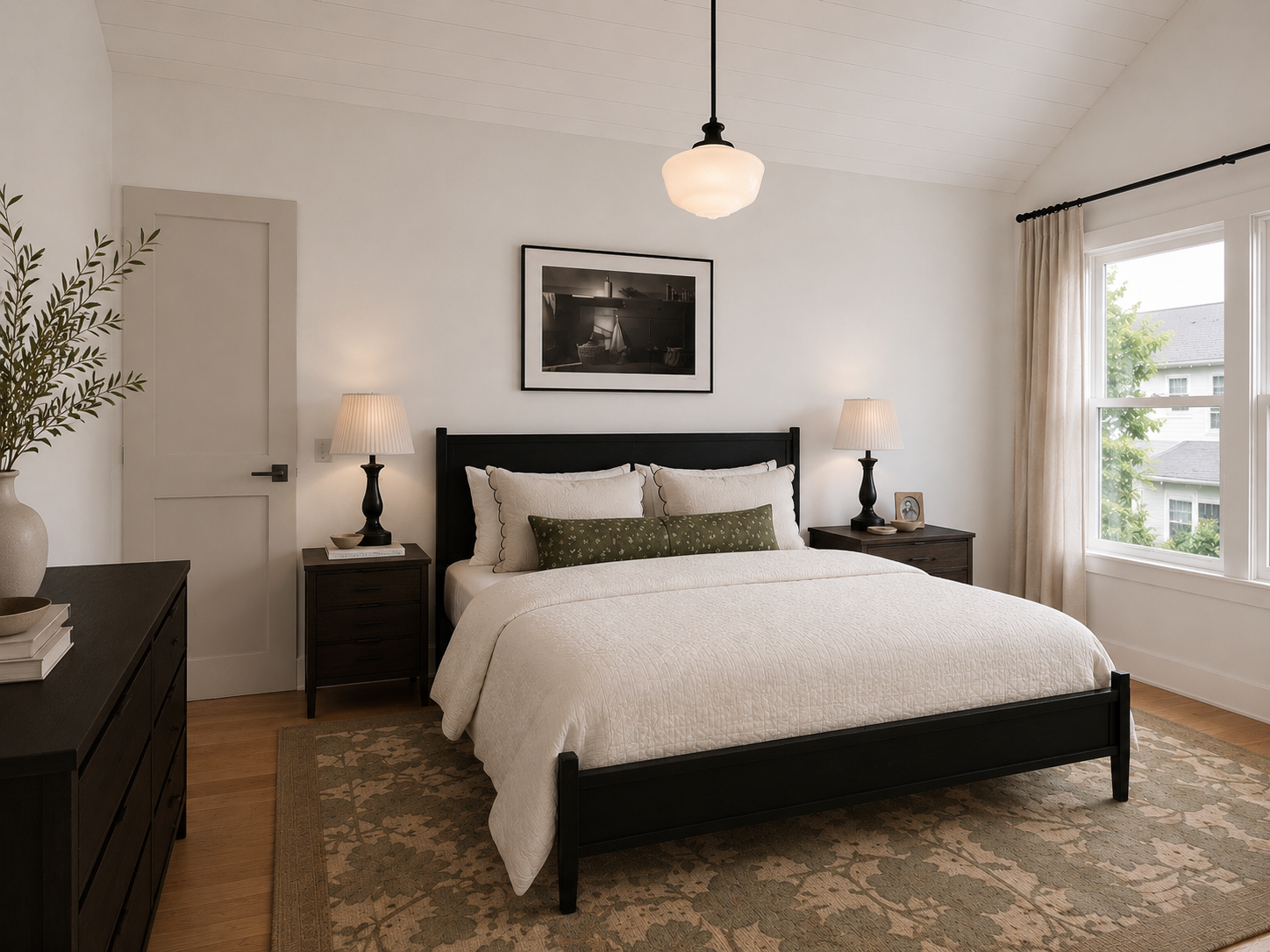 A cozy bedroom with a black bed frame, white bedding, and a green decorative pillow. Two matching nightstands with lamps flank the bed. A framed black-and-white photo hangs above the bed. Large window with beige curtains lets in natural light. Wooden dresser with a plant and some stacked books is on the left.