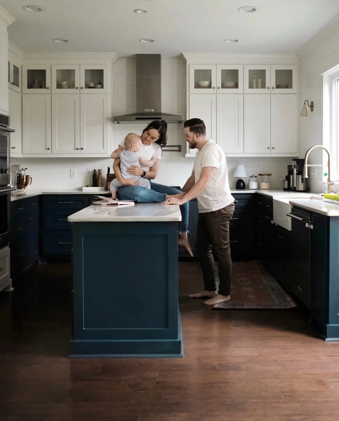 A family in a modern kitchen, with a woman holding a baby sitting on the kitchen counter, a man standing barefoot in front of her, and smiling.
