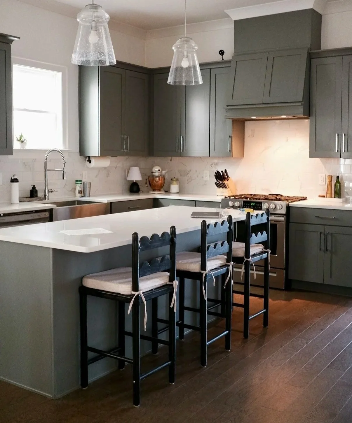 A kitchen with sage green cabinets, white countertops, beige barstools, pendant lights, and decorative elements like plants and wooden accents.