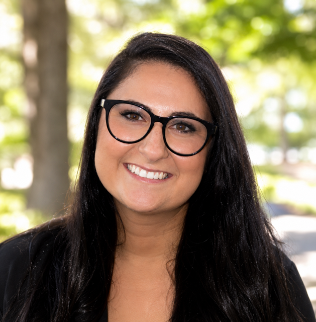 A woman with long dark hair and glasses smiling outdoors with trees and sunlight in the background.