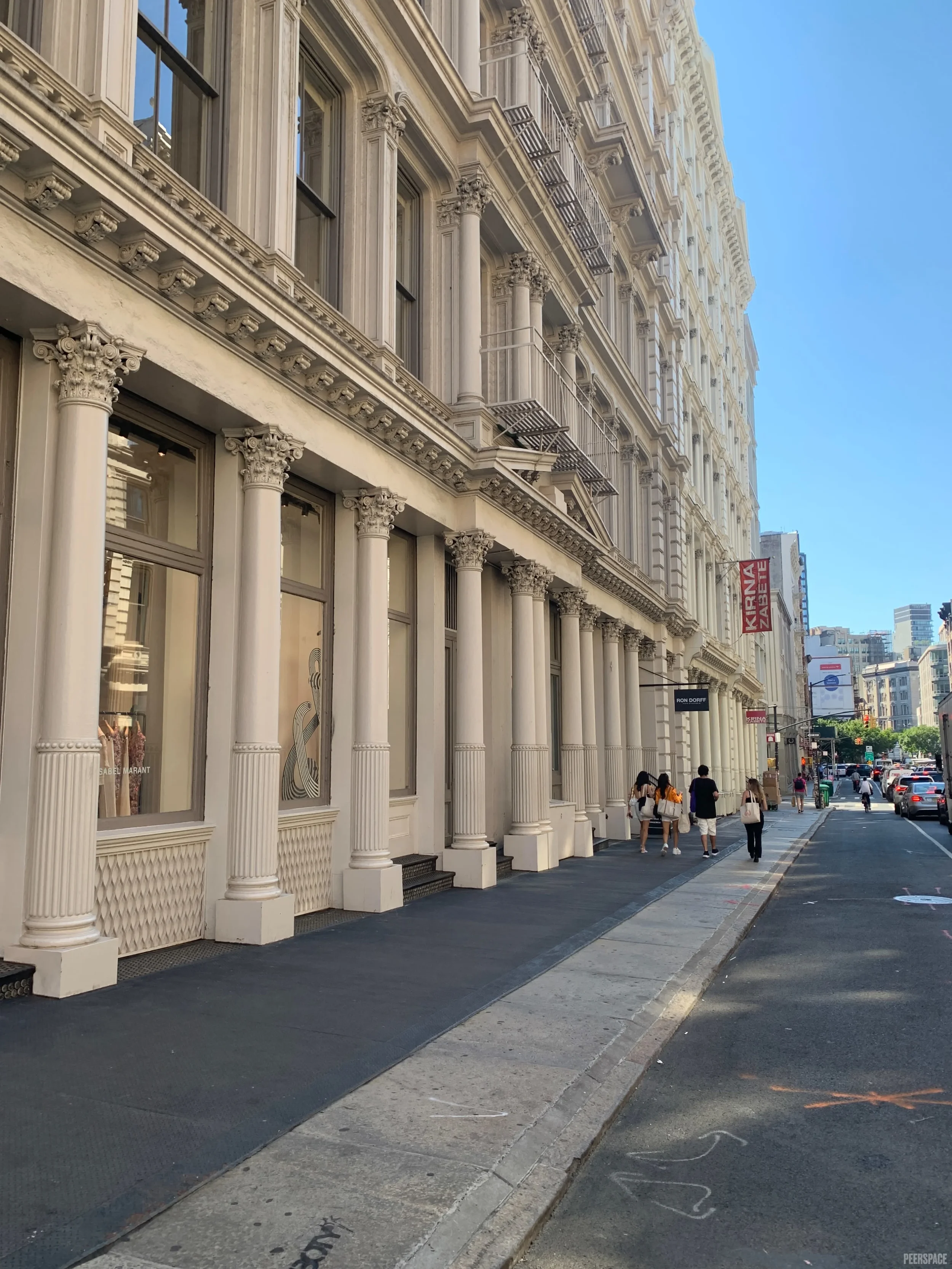 Street view of a colonial-style building with large columns, decorative details, and multiple windows; pedestrians walking on the sidewalk; clear blue sky.