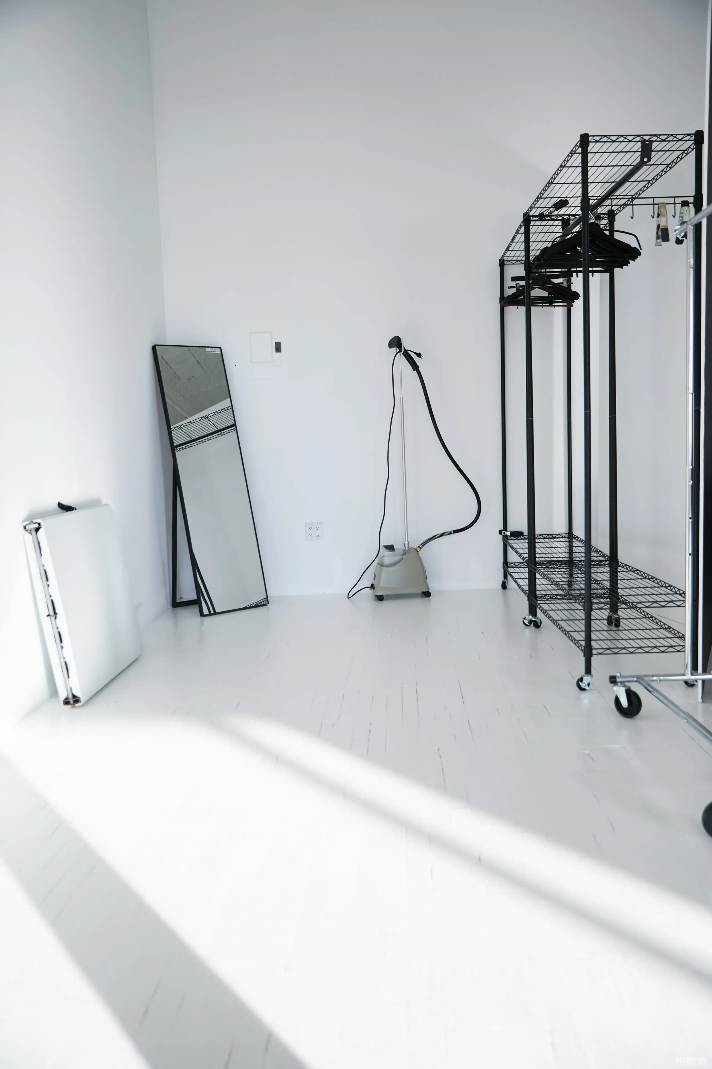 Minimalist white room with a full-length mirror, a clothes rack with hangers, a steam cleaner, and a rolling cart. Sunlight creates shadows on the floor.