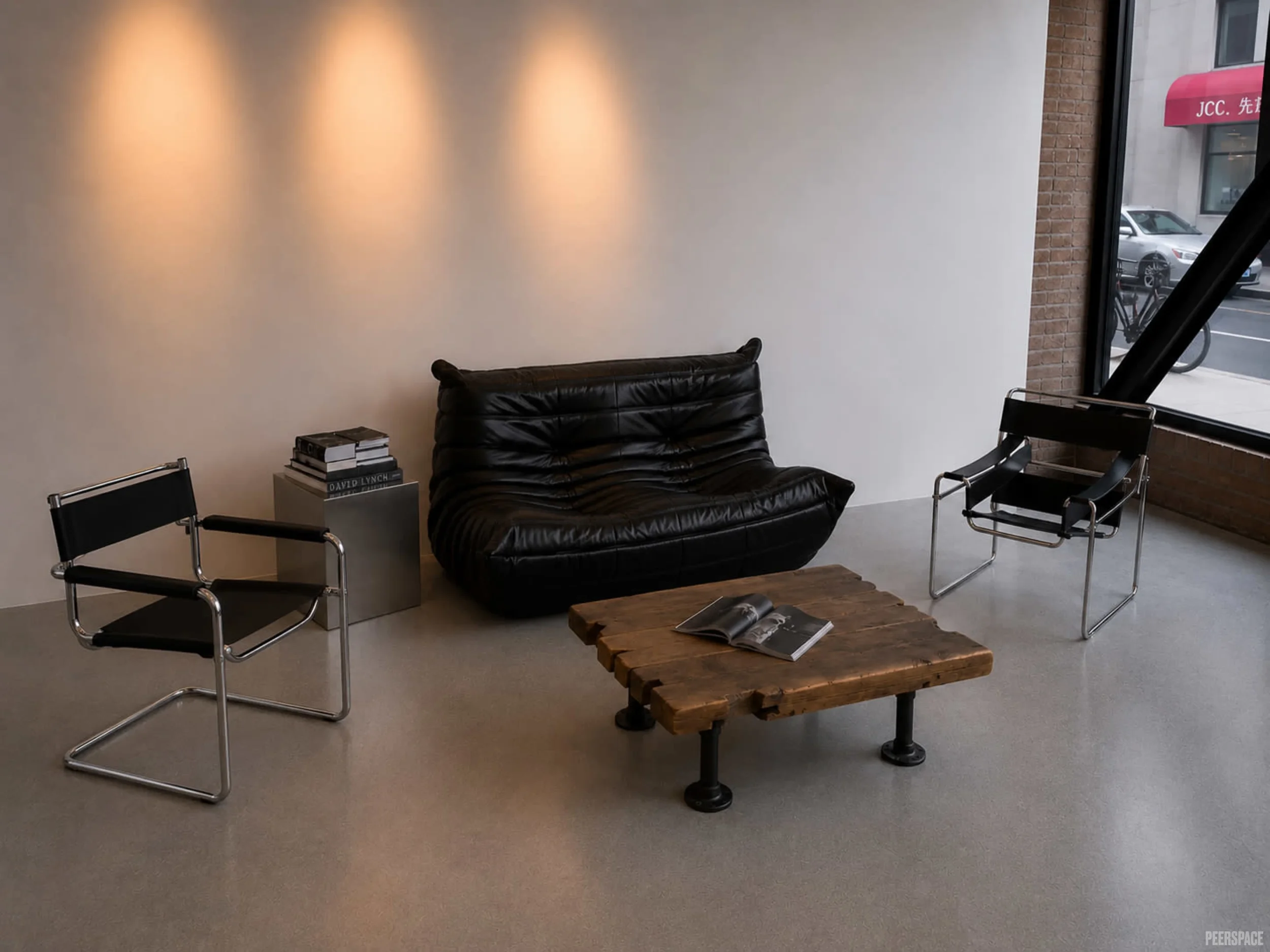 A waiting area with a black leather couch, two black metal chairs with black cushions, a stack of books on a small table, and a wooden coffee table with open magazines. There is a large window showing parked cars outside and brick wall.