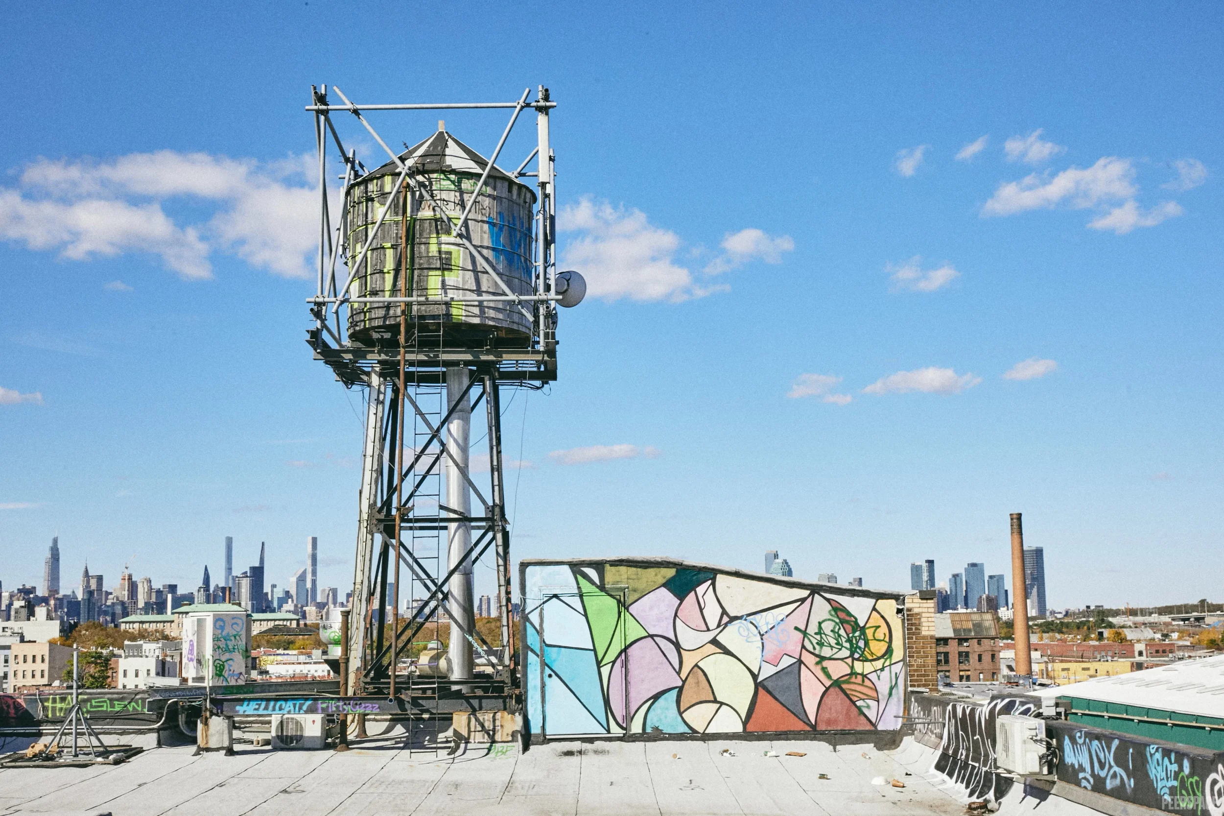 Urban rooftop with a large metal water tower covered in graffiti, surrounded by colorful street art murals, with a city skyline in the background under a clear blue sky.