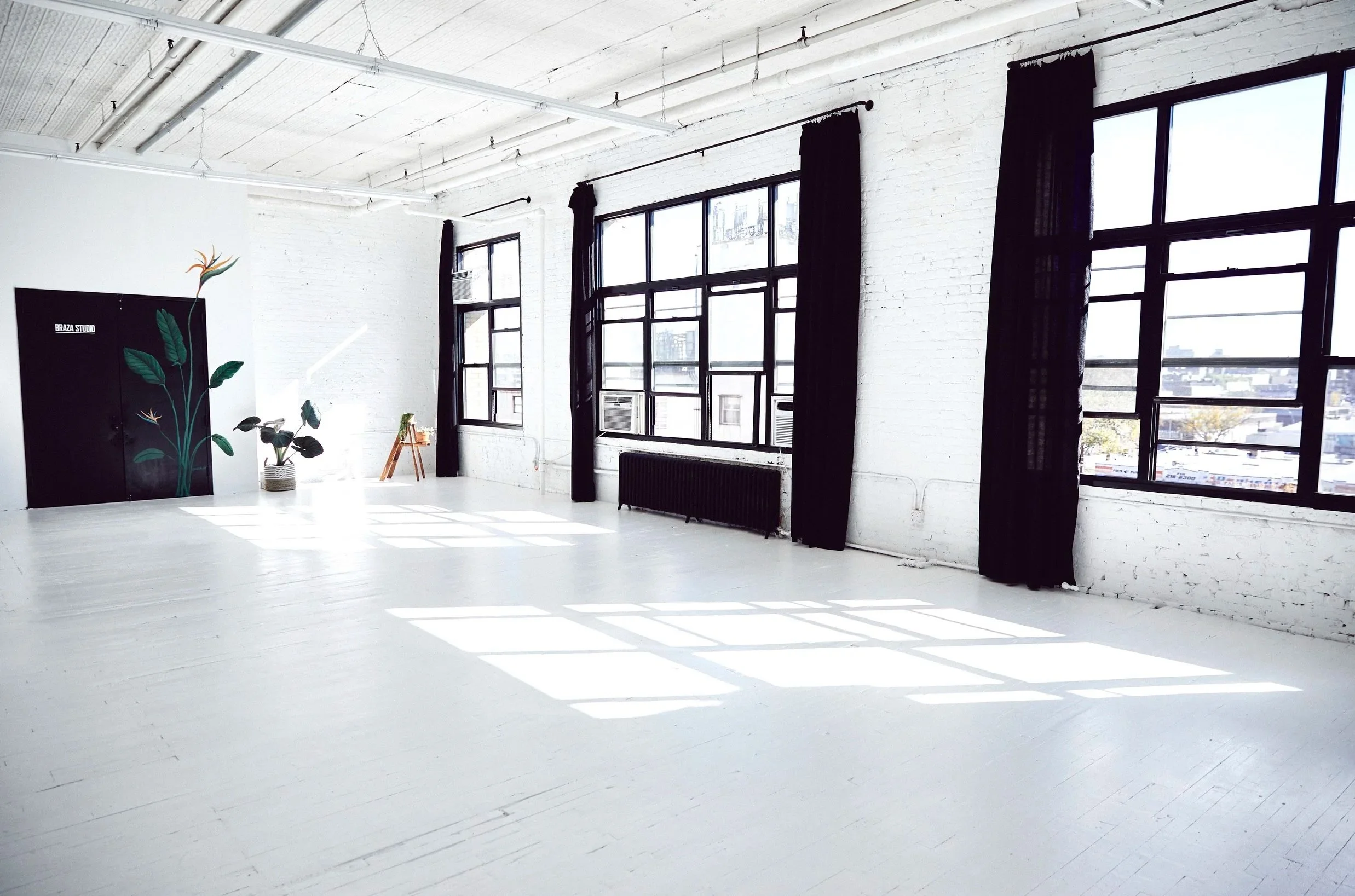 Bright, spacious room with large windows, white walls, and a white ceiling. The room has black curtains hanging by the windows, an air conditioning unit, a black cabinet with floral artwork, a potted plant, and a wooden chair. Sunlight streams into the room, creating patterns on the white floor.