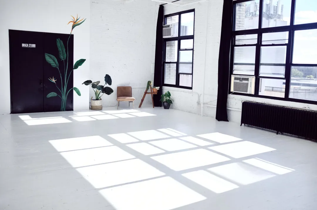 Bright, spacious studio with large windows casting shadows on a white floor, featuring a black wall with a botanical mural, potted plants, a small chair, and a window air conditioning unit.