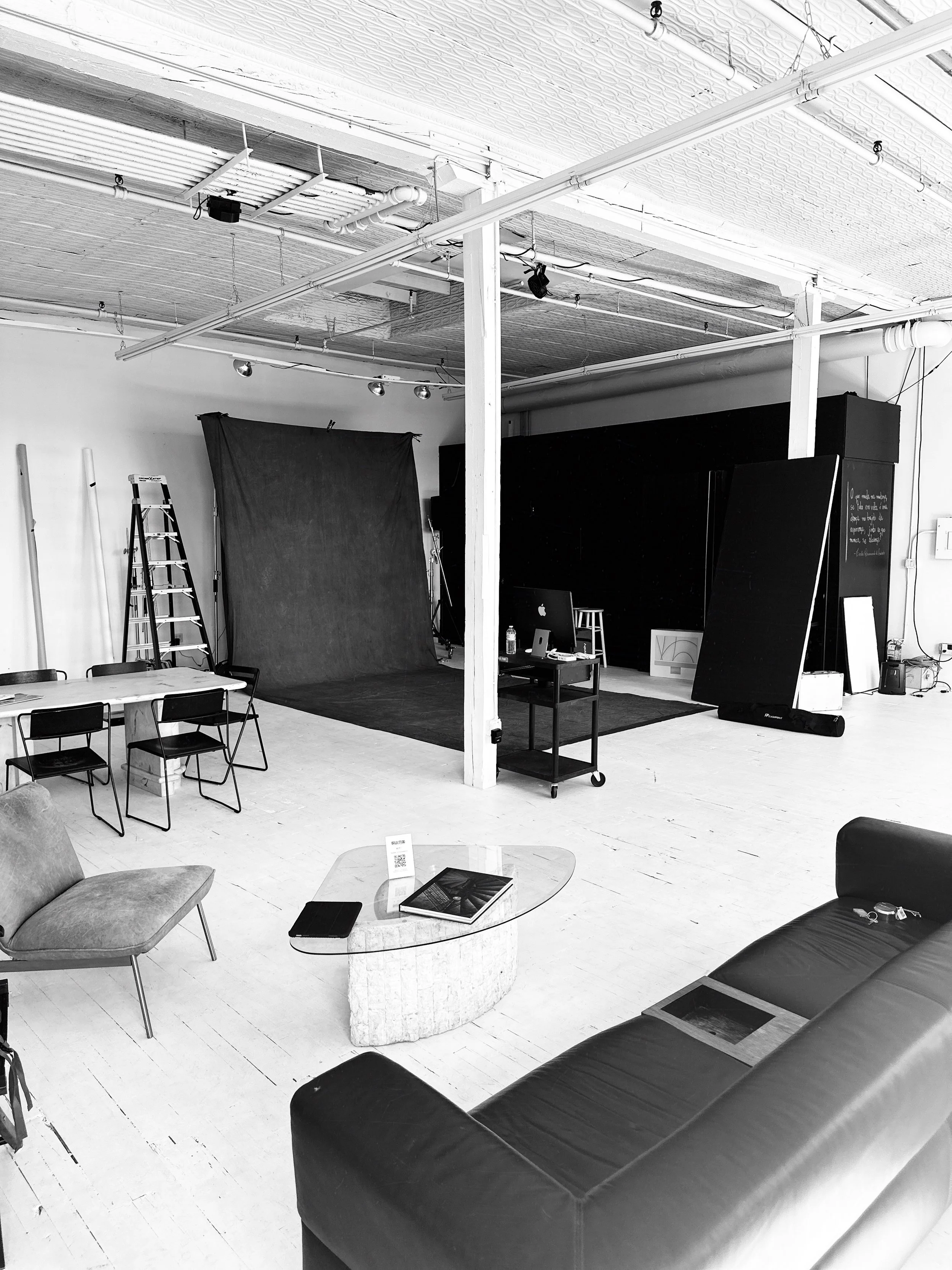 An empty photography or video studio with a black backdrop set up, various chairs, a ladder, and a table with a book and a cellphone. The ceiling has exposed pipes and lighting fixtures.