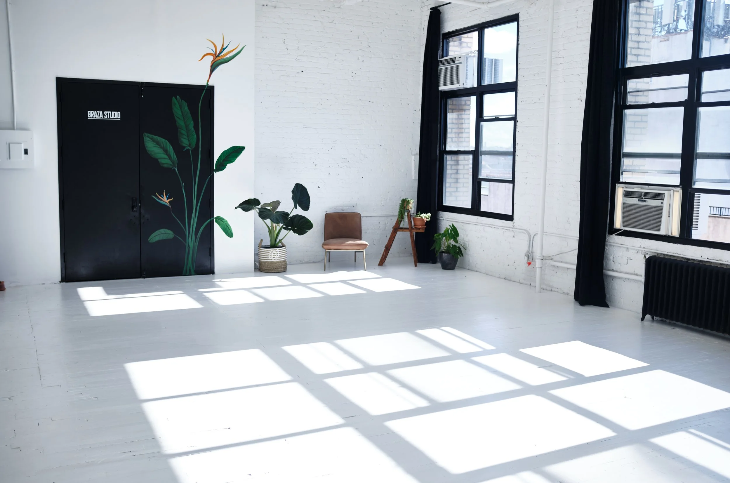 Bright, minimalist room with white walls and floors, large black-framed windows letting in natural light, some potted plants, a brown chair, black curtains, and a black cabinet with a painted floral design.