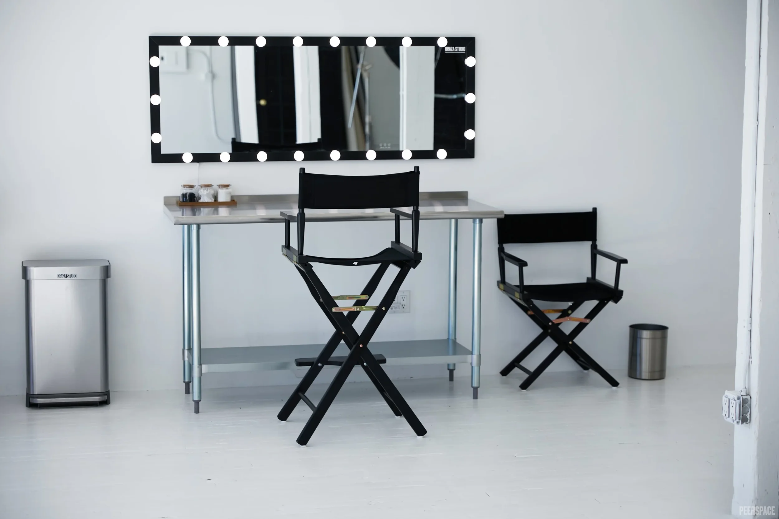 Empty makeup or dressing room with a large mirror surrounded by bright bulbs, a side table with small jars, two black folding chairs, a trash can, and electrical outlets on the wall.