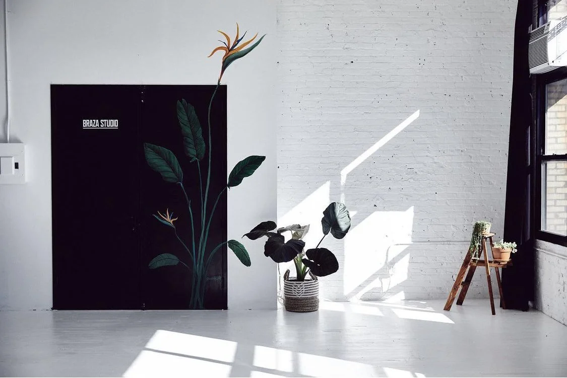 Interior room with white brick wall and large window letting in natural light. There is a large black painting or door with a floral design, two potted plants, and a small wooden table with another potted plant on it.
