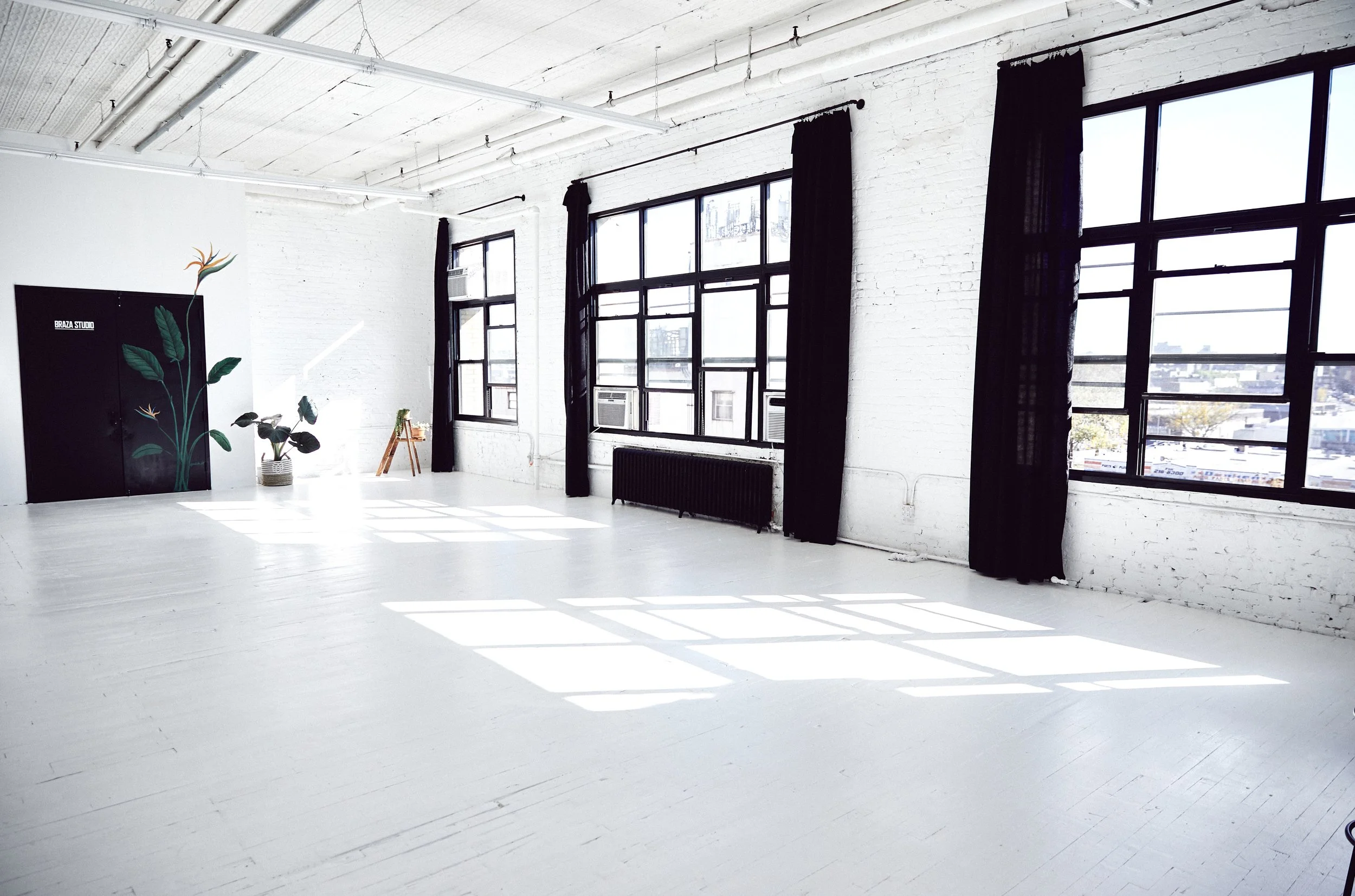 Brightly lit room with large windows, white brick walls, black curtains, and a white painted wooden floor. There are plants, a black cabinet with painted leaves, and some small decorative items.
