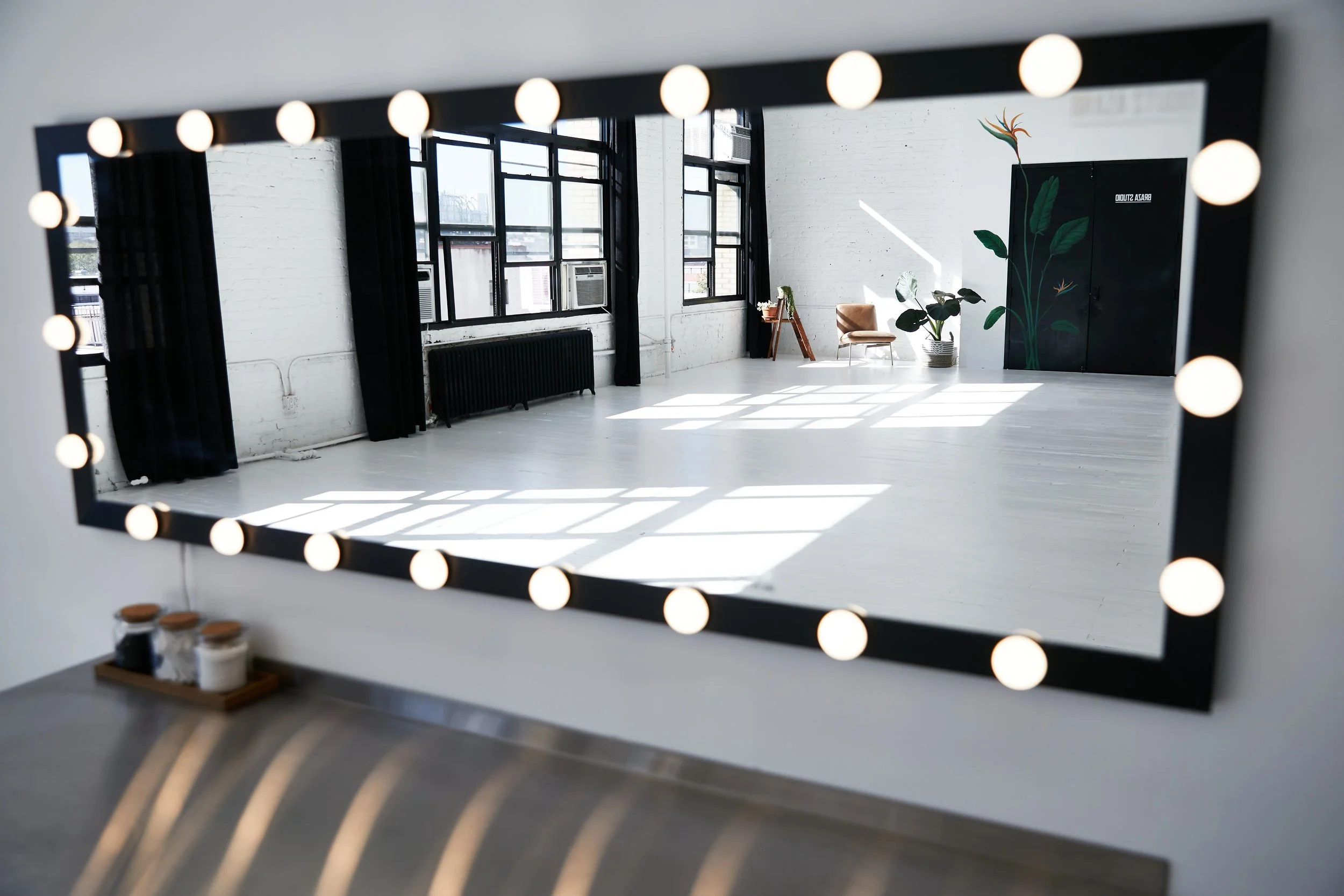 A mirror with a black frame and round illuminated bulbs on a wall, reflecting a bright room with large windows, black curtains, a brown chair, potted plant, and a black cabinet with green leaf design.