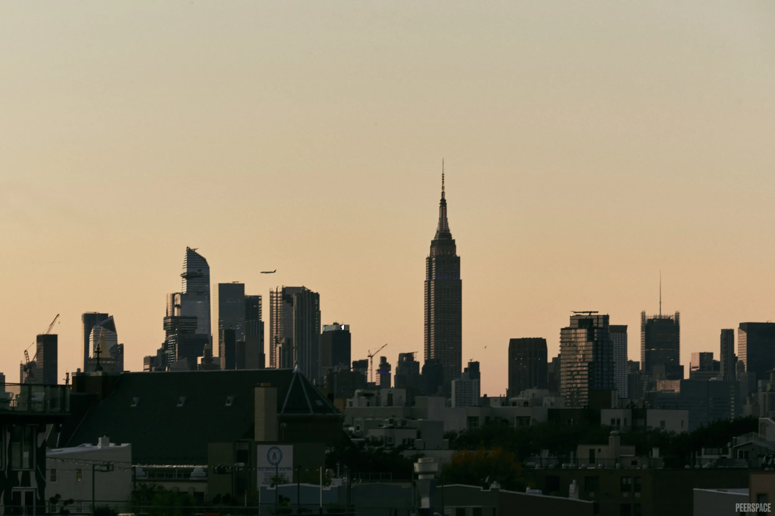 City skyline during sunset with various skyscrapers, including the Empire State Building, and an airplane flying in the sky.